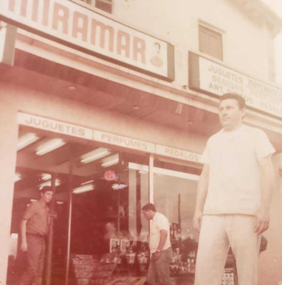 Three young men standing in front of a store with signs for snacks, perfumes, and cigarettes, with the store's storefront visible.