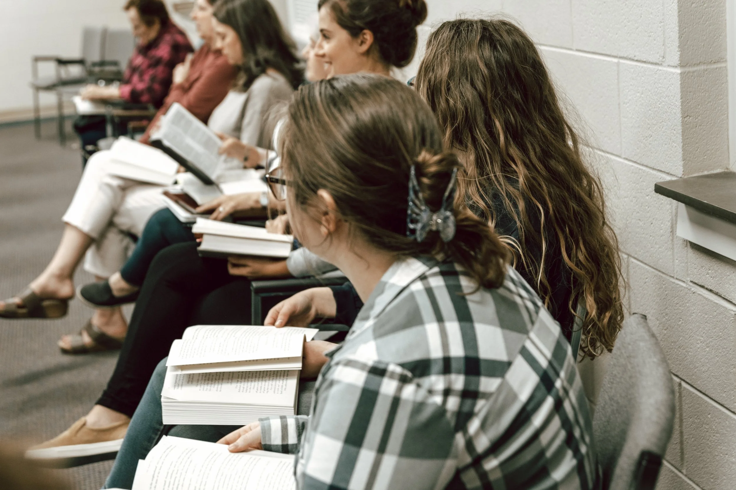 A group of women sitting on chairs in a classroom or conference room, reading books or listening attentively.