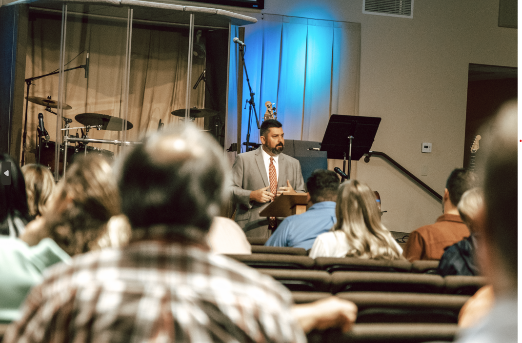 A man in a gray suit giving a speech at a church or conference event, standing at a podium with musical instruments and microphones on stage behind him, audience seated in front.