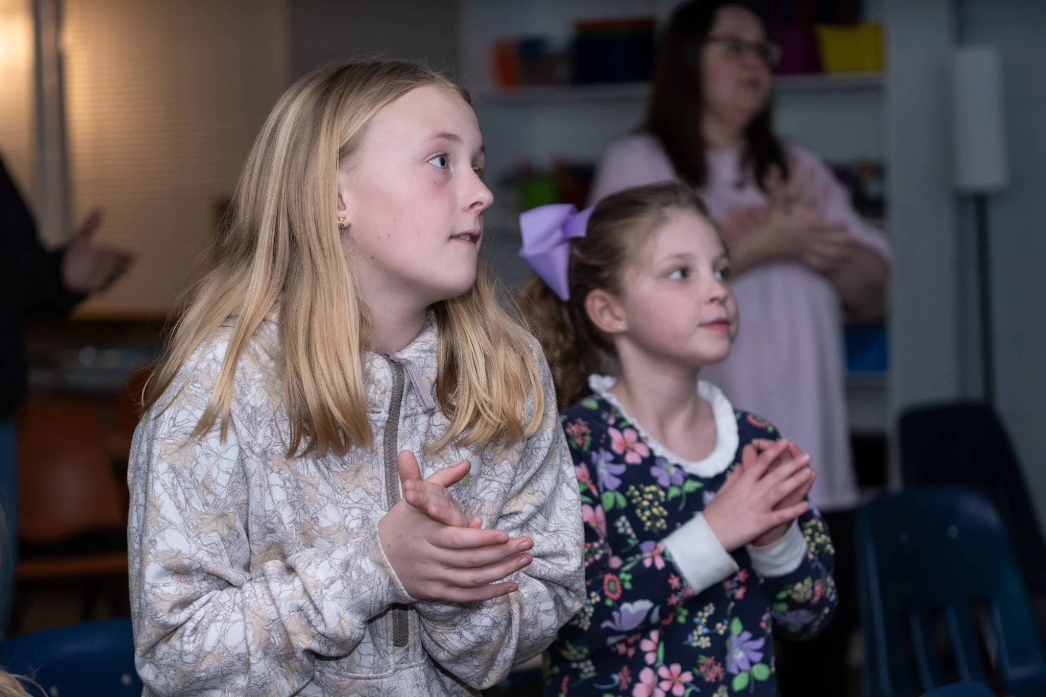 Two young girls are standing with their hands clasped, watching something with expressions of interest. A woman stands in the background with her hands folded.