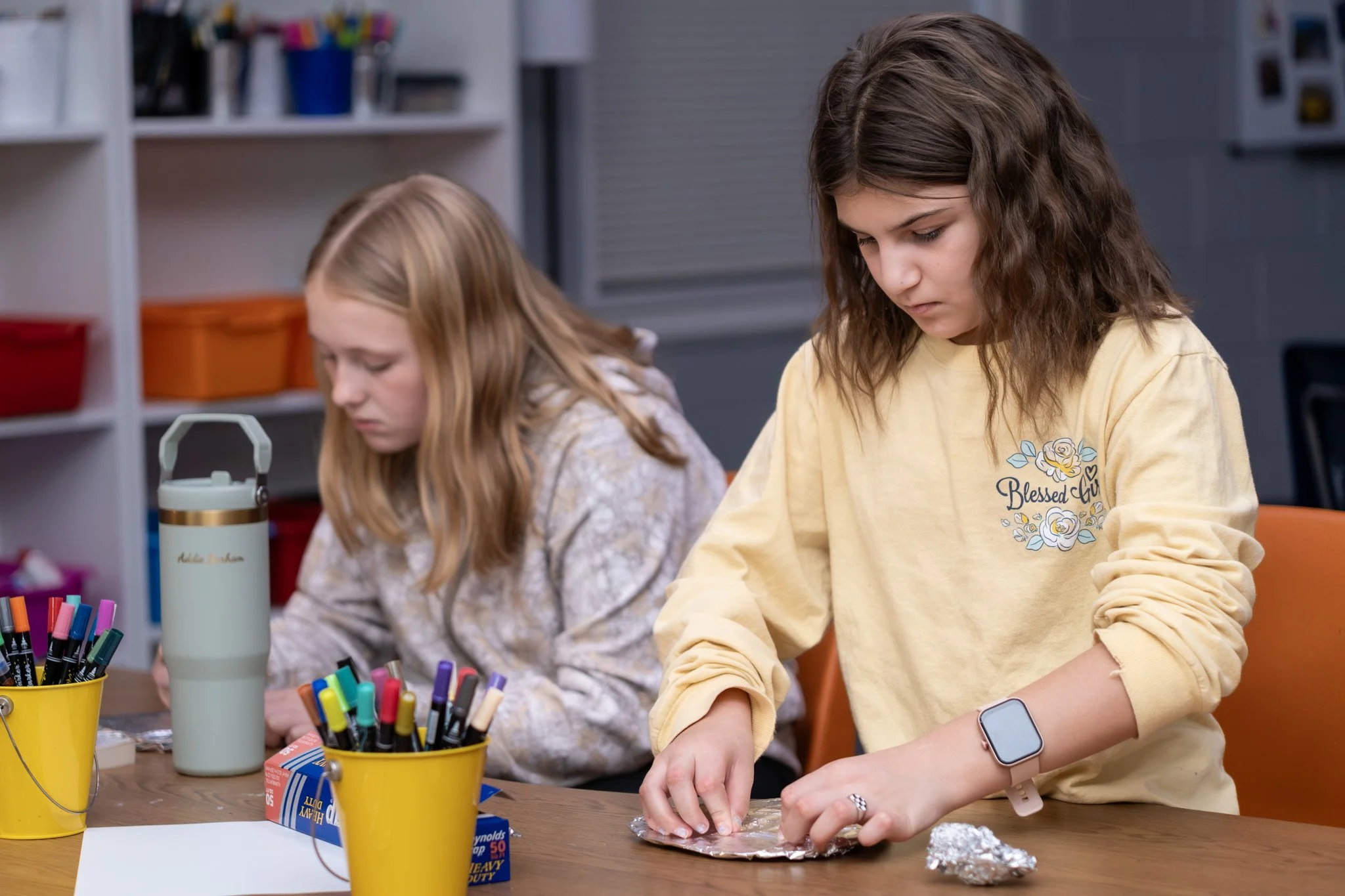 Two young girls sitting at a table engaged in a craft activity. One girl in a yellow sweatshirt is handling aluminum foil, while the other girl in a patterned hoodie looks downward. The table has colorful markers, a tension thermal flask, and some craft supplies.