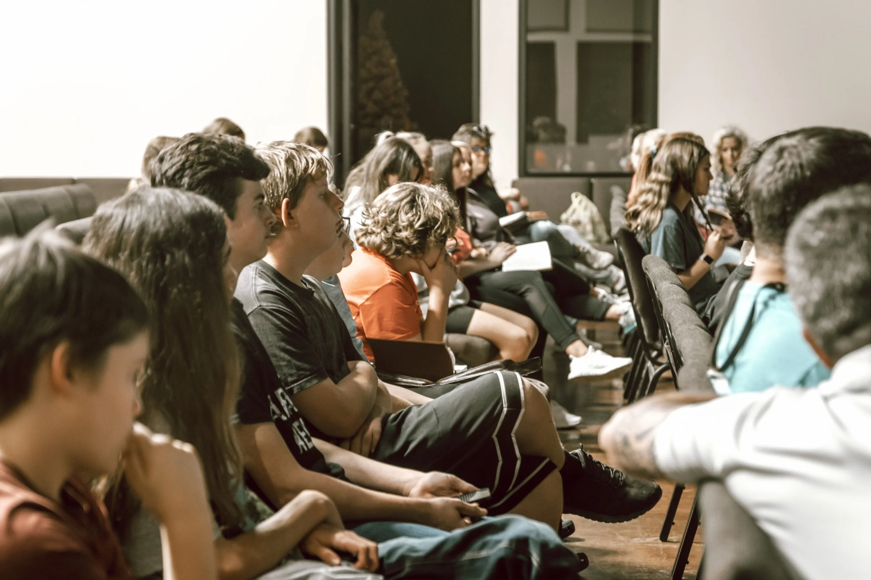 Young people sitting in a room, attentively listening to a speaker or presentation, some taking notes, with a window in the background.