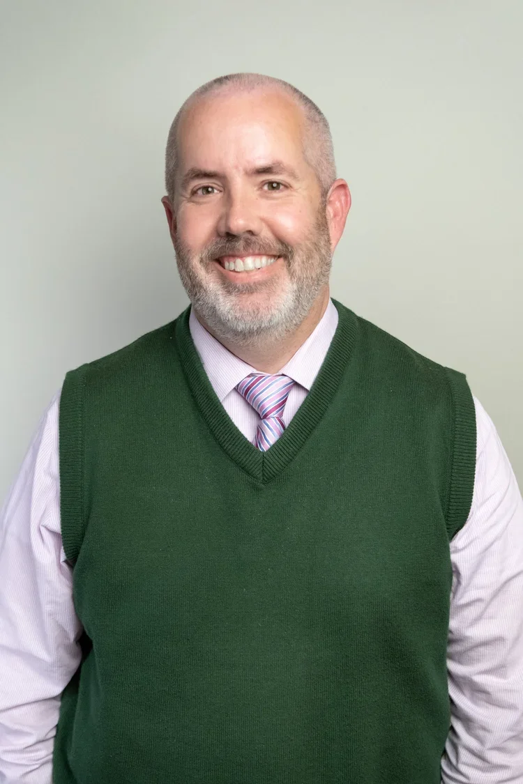 A smiling man with a beard and short hair, wearing a white dress shirt, a striped pink and blue tie, and a green sleeveless sweater, standing against a plain light-colored background.