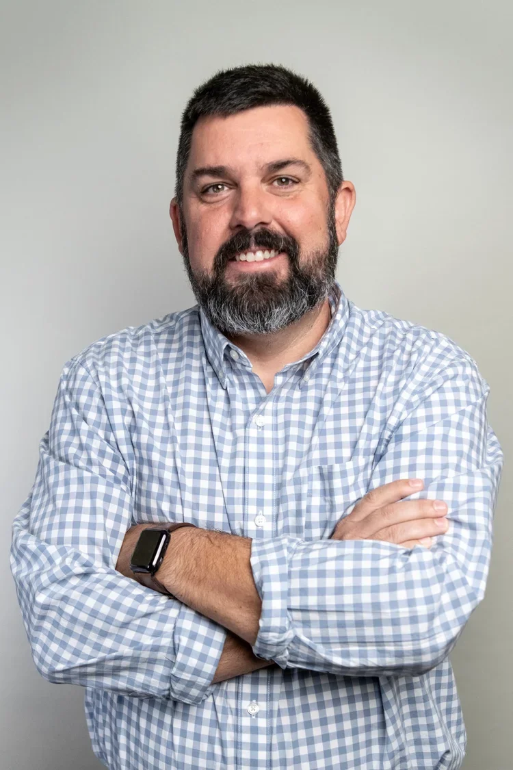 Man with a beard wearing a checkered button-up shirt and a smartwatch, standing against a plain light background.