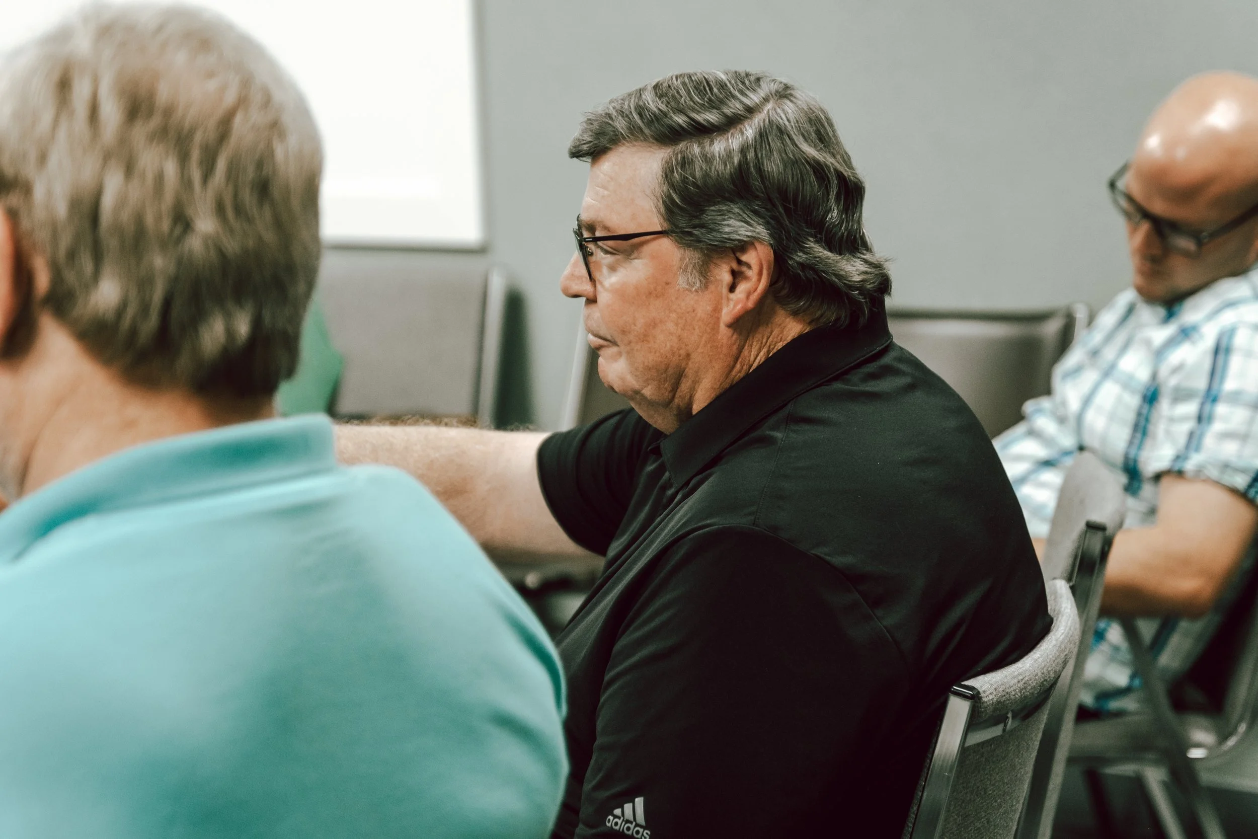 Group of people sitting in a room, with a focus on a man with glasses and dark hair wearing a black shirt, appear to be attending a meeting or seminar.