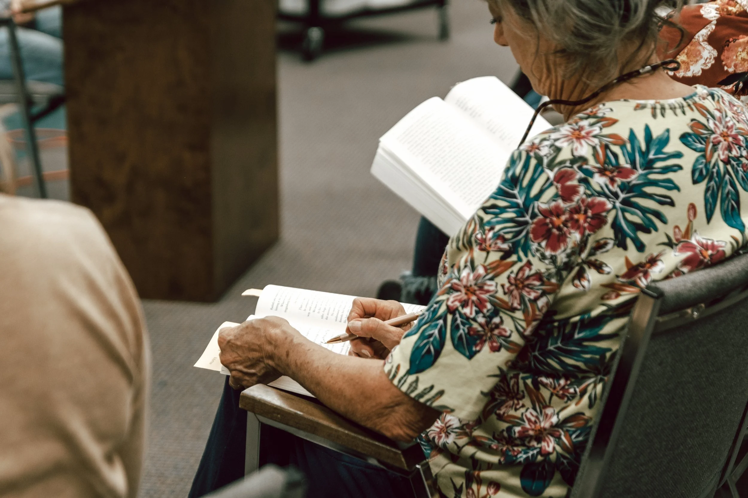 An elderly woman with gray hair and glasses, wearing a floral patterned shirt, sitting in a chair, reading a book, and writing in a notebook with a pen.
