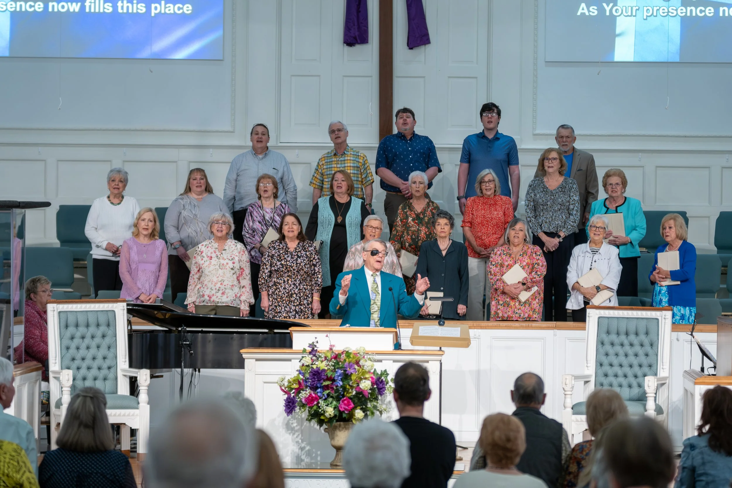 A choir of diverse adults standing on stage inside a church, singing, with a conductor in front gesturing, audience seated in front, and a large flower arrangement on the pulpit.