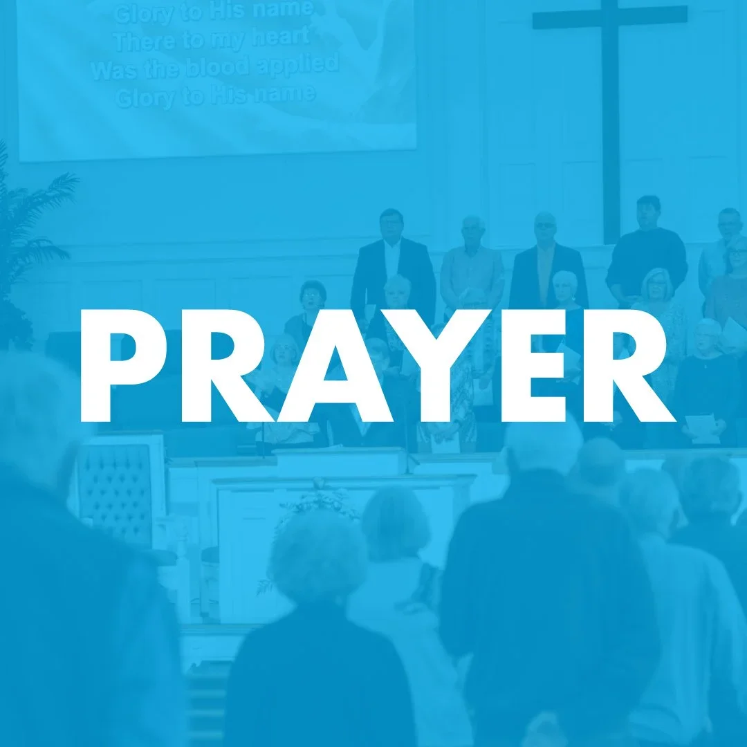 People standing and sitting in a church during prayer with a large cross on the wall and a screen displaying song lyrics in the background.