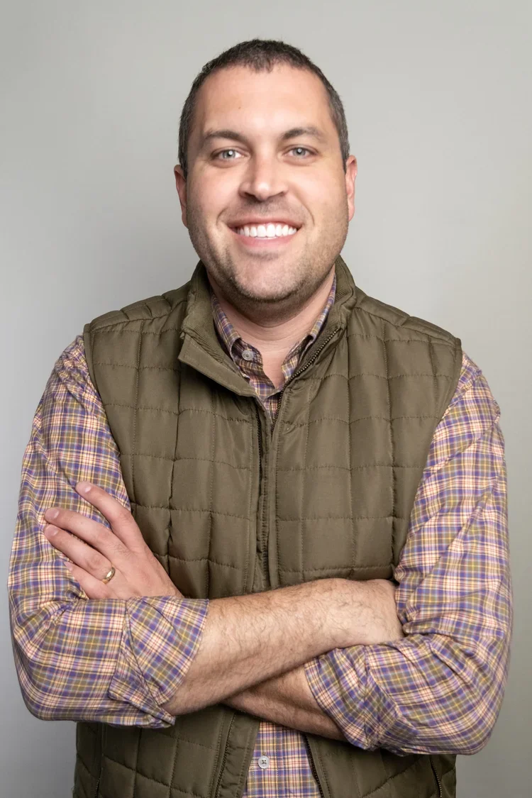 A smiling man with short brown hair, wearing a plaid shirt and a green quilted vest, standing against a plain gray background.