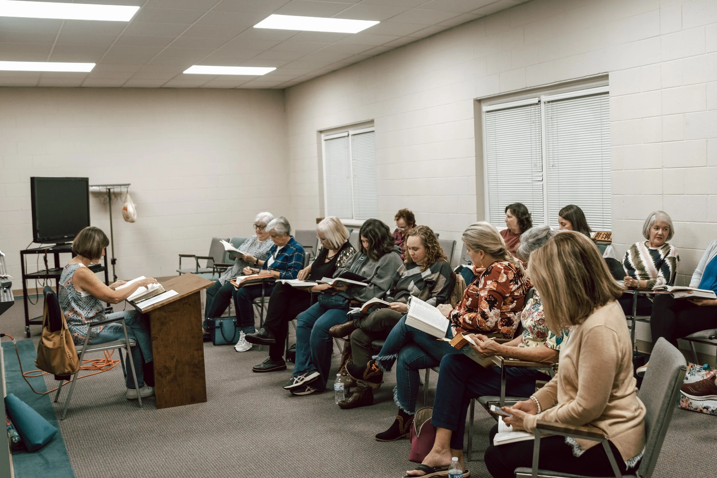 A group of women sitting in chairs in a classroom or seminar room, reading books or notes, listening to a woman at the front, with a large monitor on a stand and a coat rack in the background.
