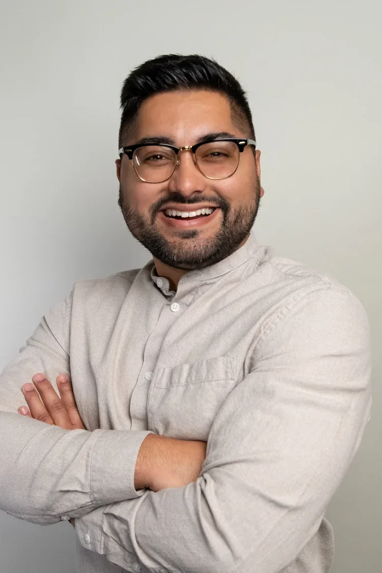 A smiling man wearing glasses and a beige button-up shirt, standing with arms crossed against a plain light-colored background.