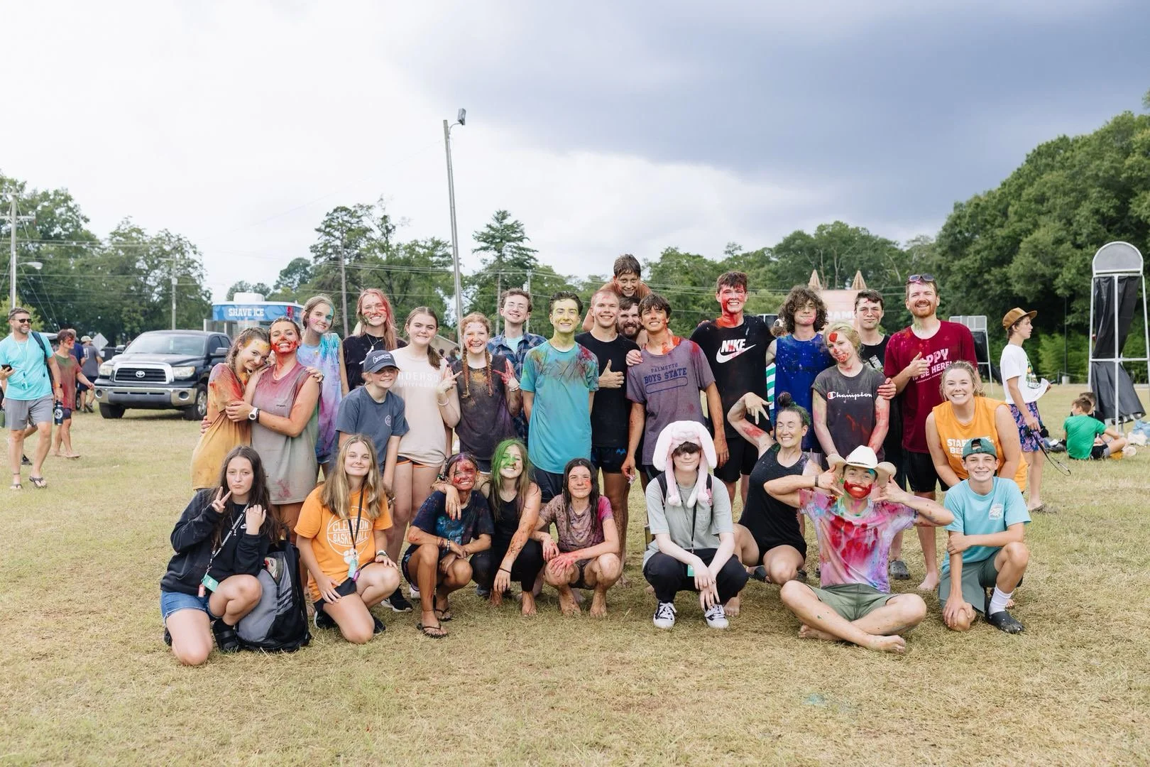 Group of children and teenagers with colorful face and body paint, some wearing casual clothing and hats, posing together outdoors on a grassy field during daytime.