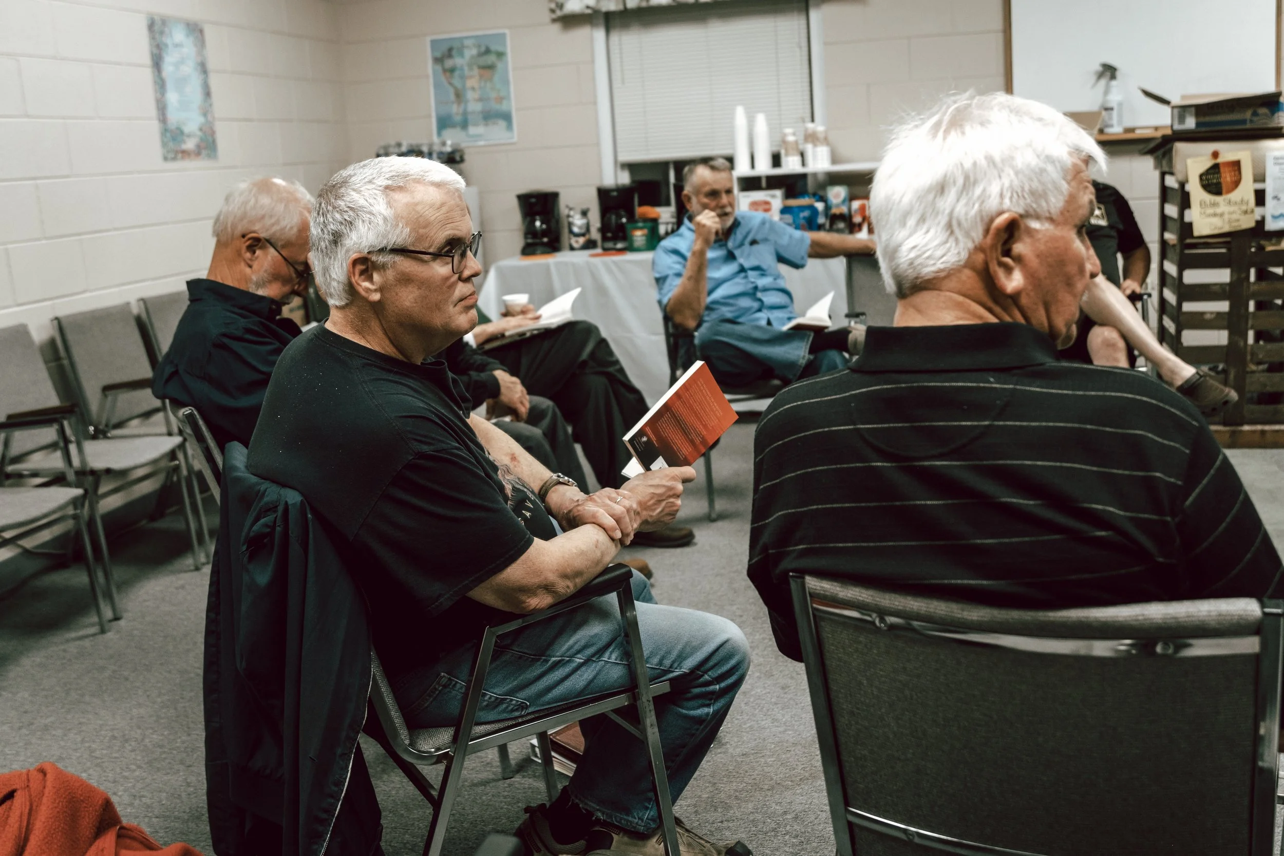 Group of older adults seated in a room, some reading pamphlets, others listening attentively, with coffee setup and maps on the wall in the background.