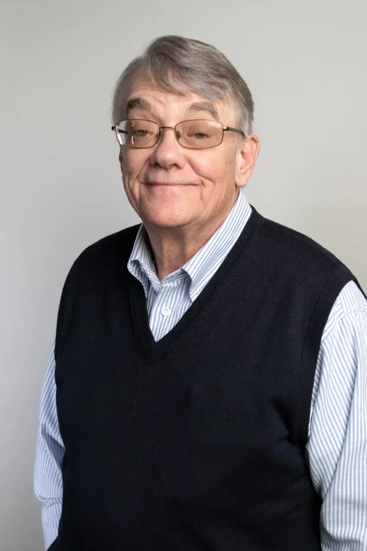Portrait of an older man with glasses, wearing a striped shirt and a dark sleeveless sweater, standing against a plain light-colored wall.