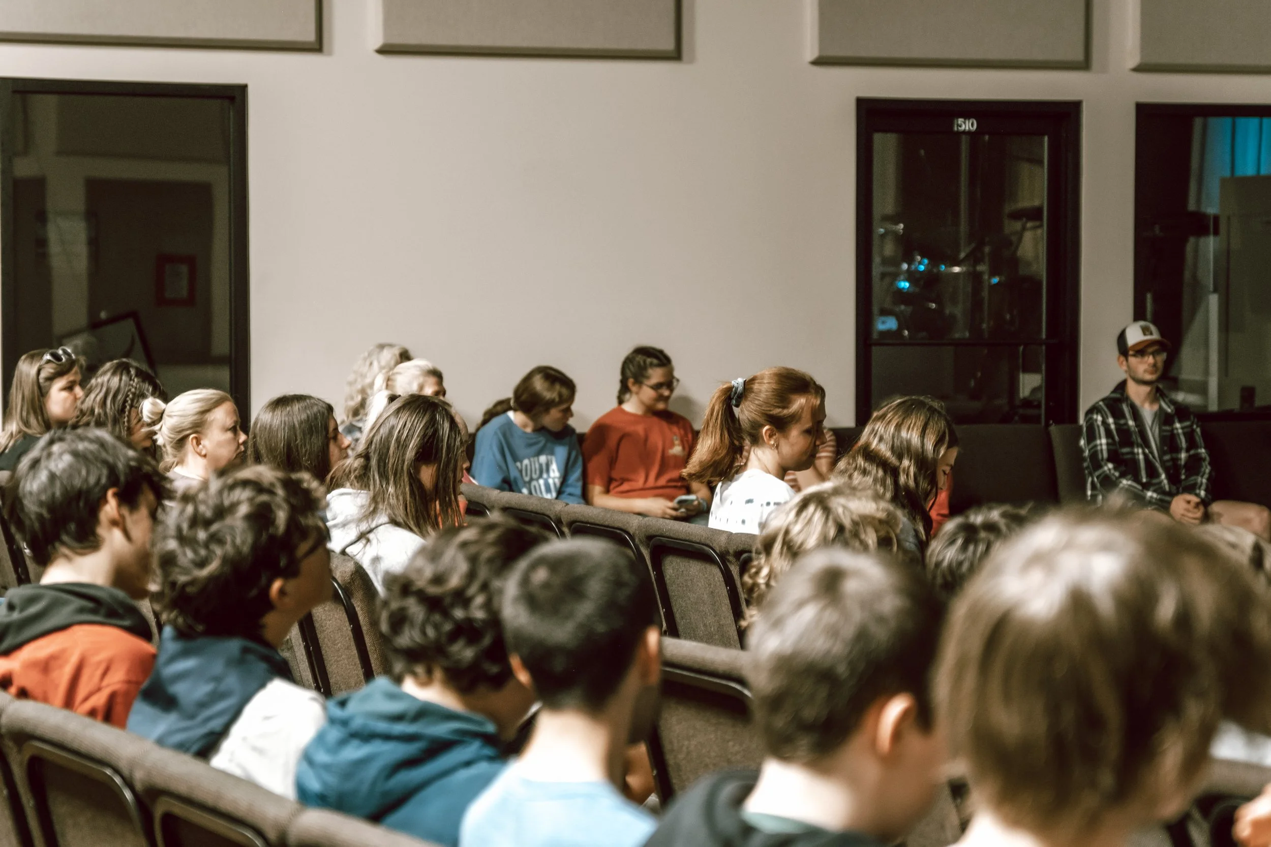 Group of people sitting in an audience in a dimly lit room, attentively listening to a presentation or speech.