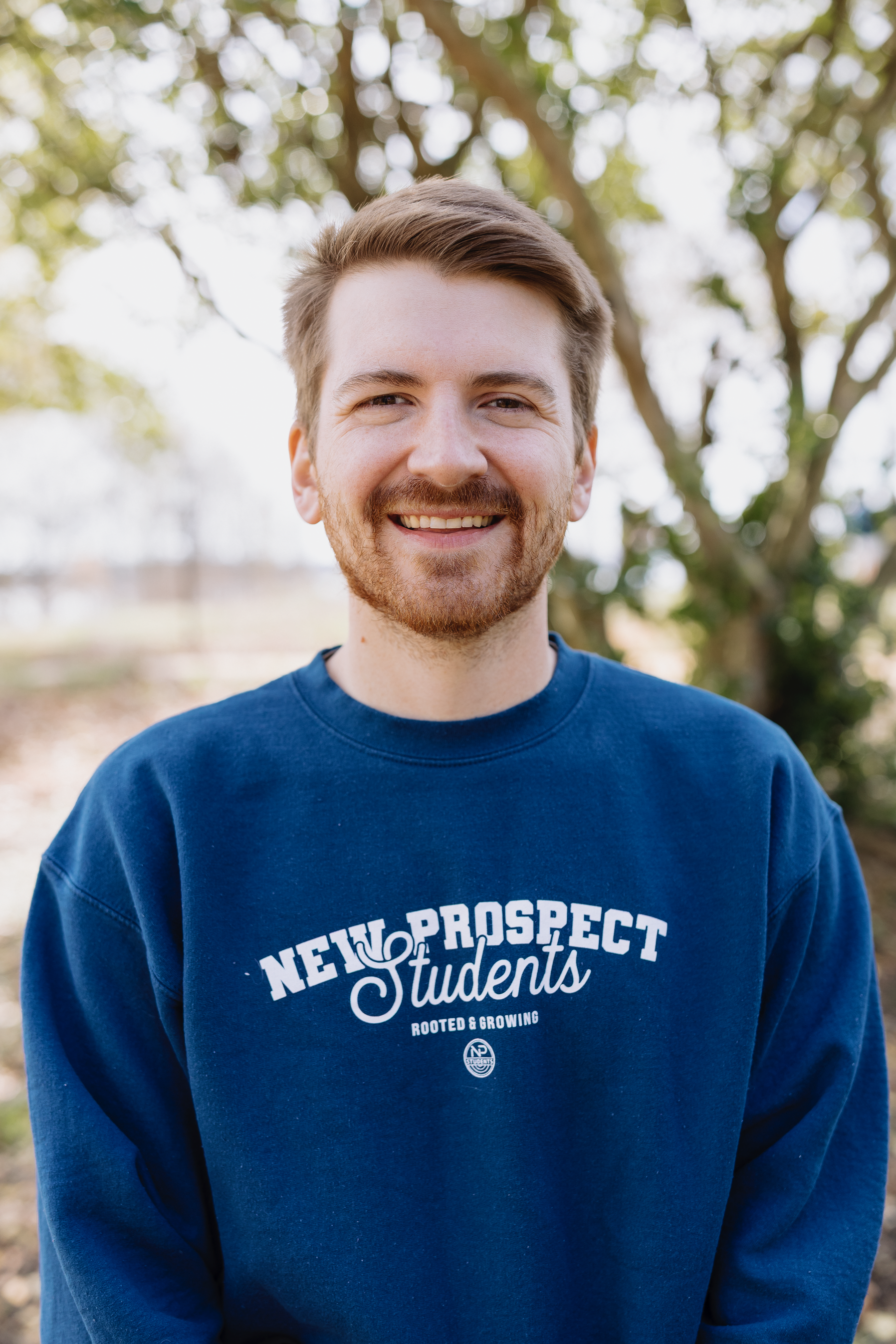 Smiling young man with brown hair and beard wearing a blue sweatshirt that says 'New Prospect Students Rooted & Growing' standing outdoors in front of a tree with green leaves.