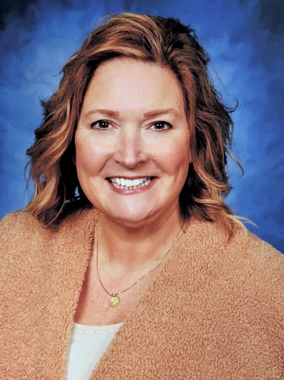 A woman with shoulder-length wavy ginger hair, wearing a beige textured jacket, white top, and a gold necklace, smiling in front of a blue background.