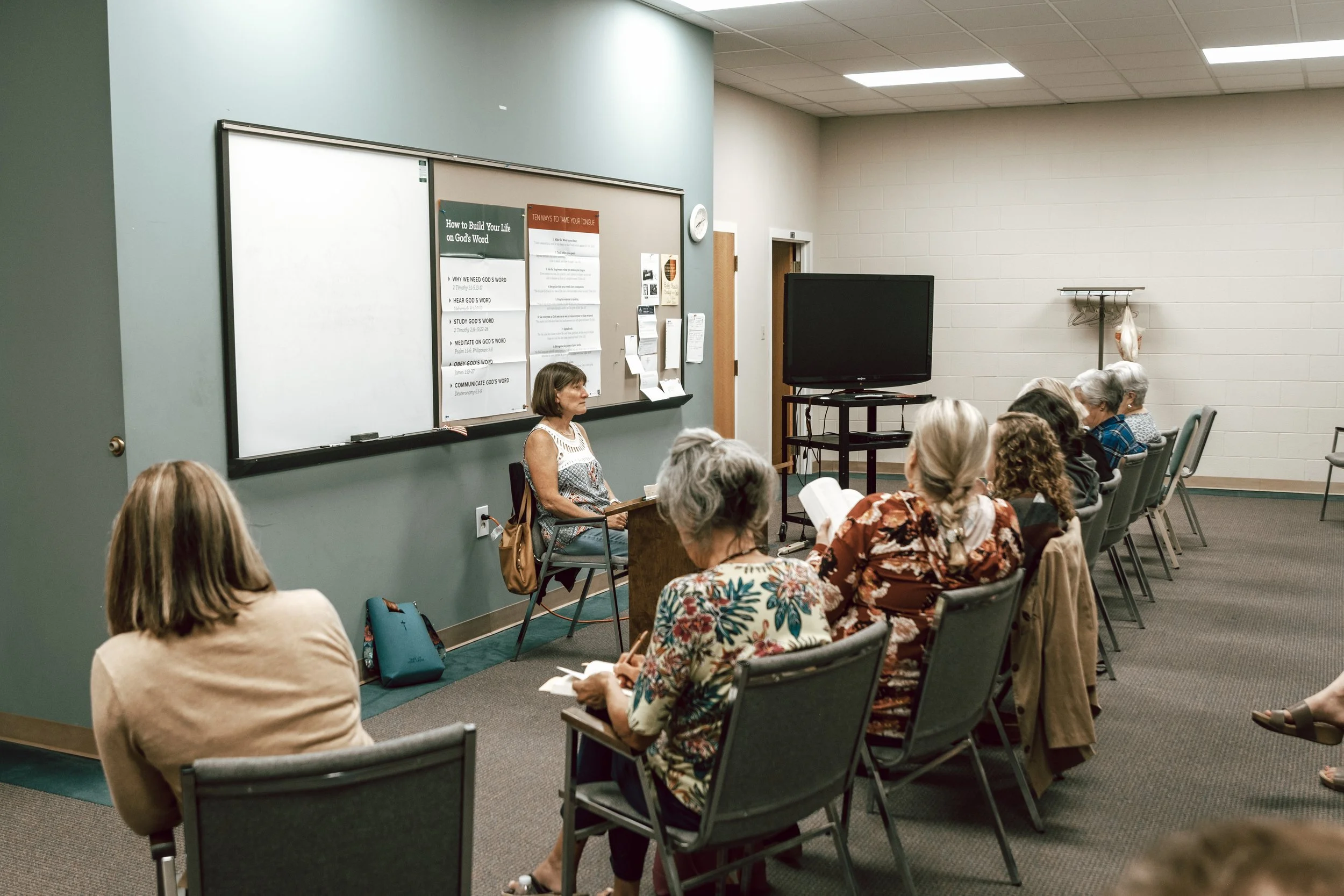 A woman giving a presentation to a group of older women seated in a classroom with a blackboard, a large television, and informational posters on the wall.