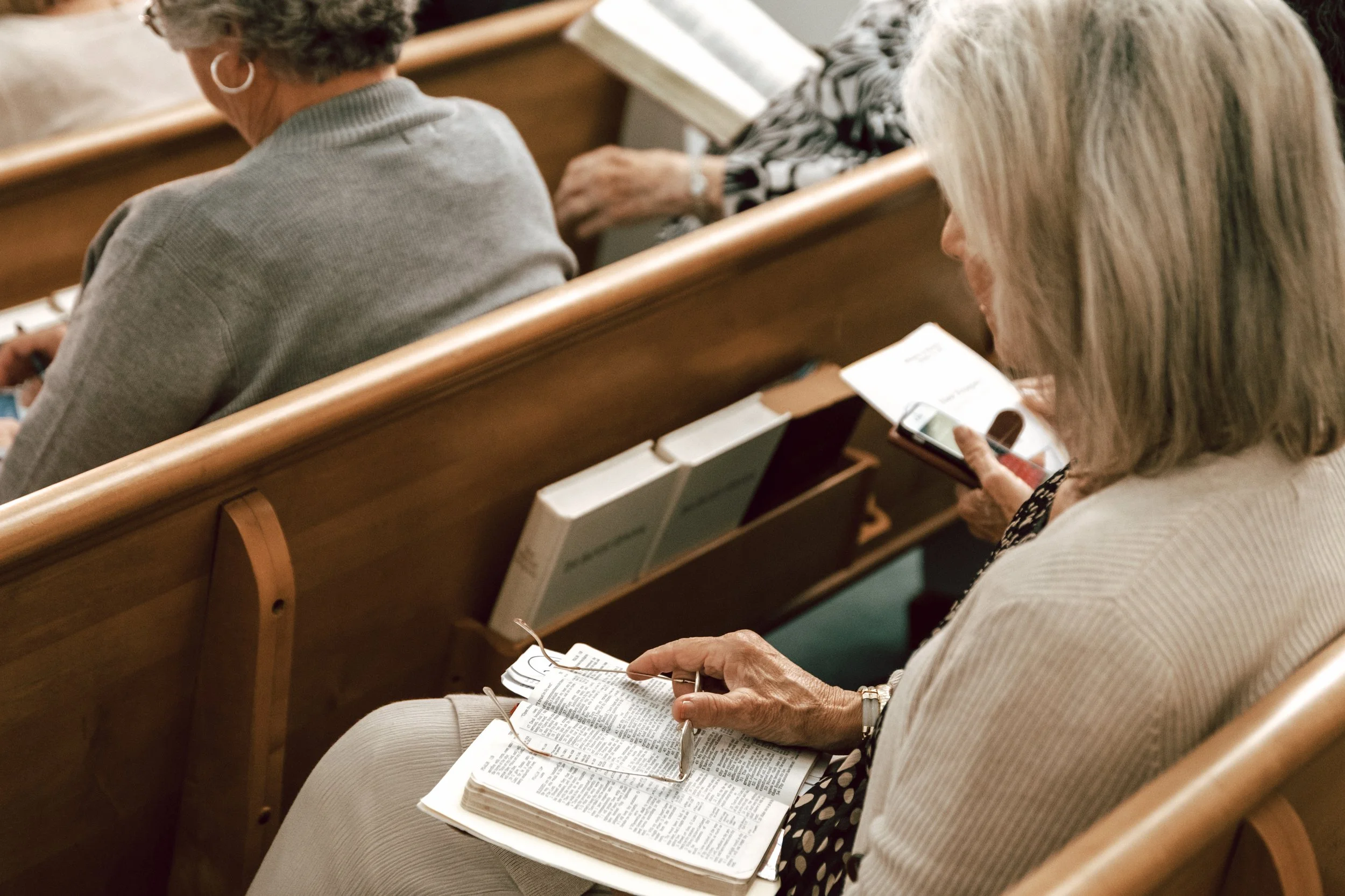 An elderly woman sitting in a church pew reading a book and holding a pair of glasses, with her phone in her other hand, beside another woman using her phone.