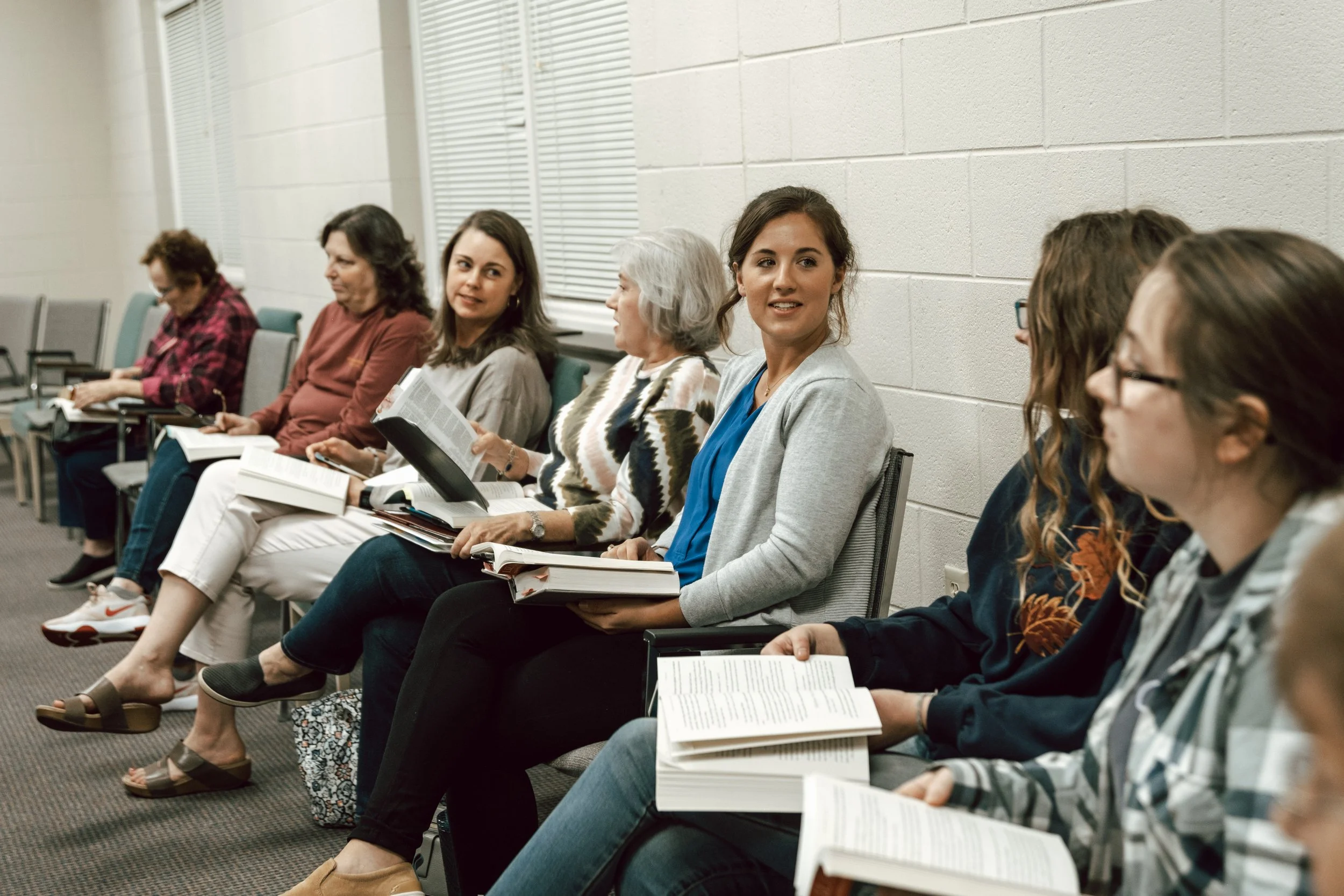 A group of women sitting on chairs, some holding books and taking notes, in a classroom or conference room setting.
