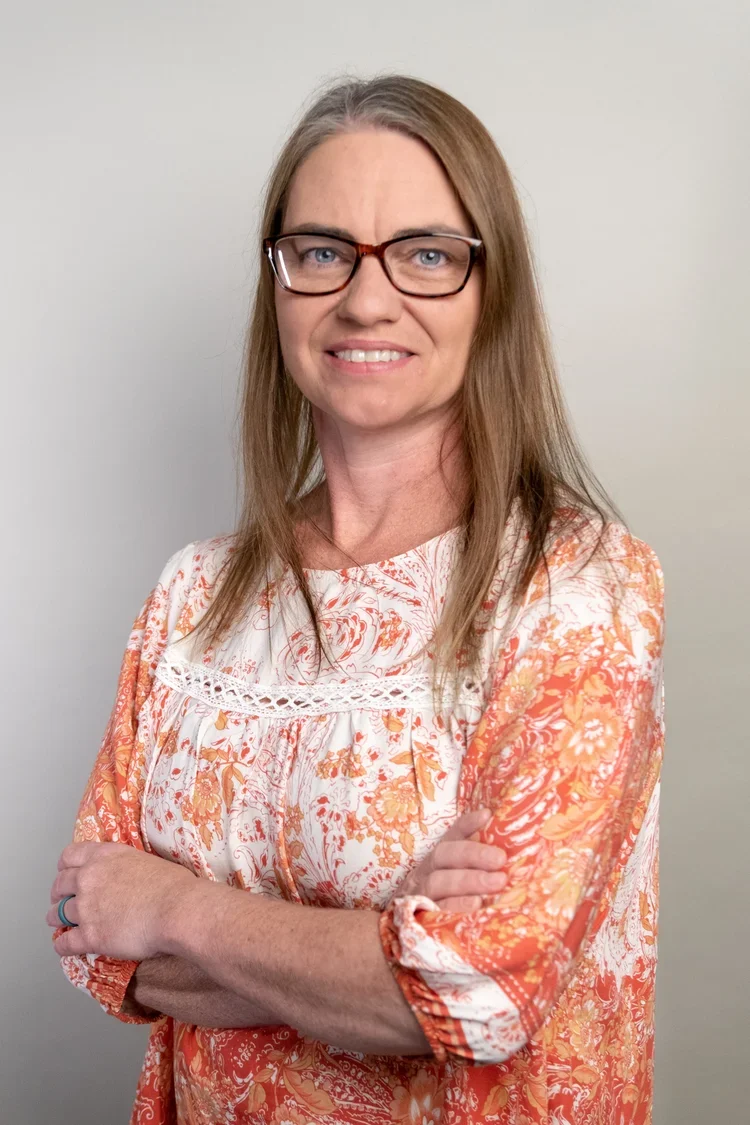 A woman with shoulder-length light brown hair, glasses, and a friendly smile, wearing a patterned blouse with orange, white, and yellow floral designs, standing against a plain light-colored wall.