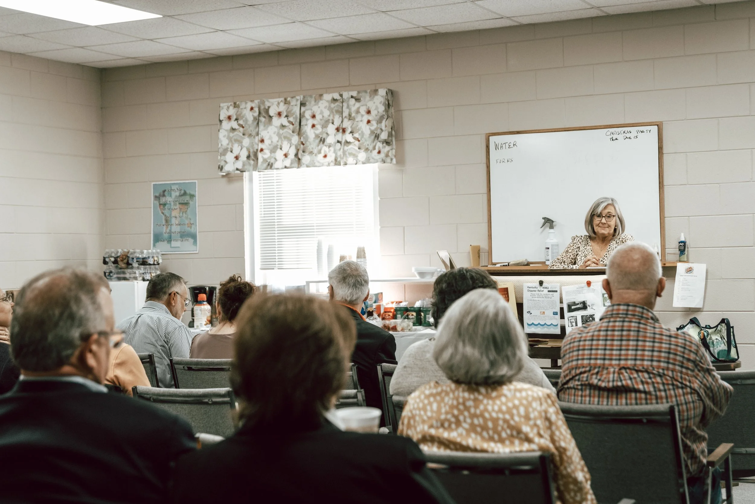 An elderly woman giving a presentation to a seated audience in a classroom or community room. The woman is standing behind a whiteboard, with notes written on it, and there are various supplies and informational materials on a table in front of her. The audience is listening attentively.
