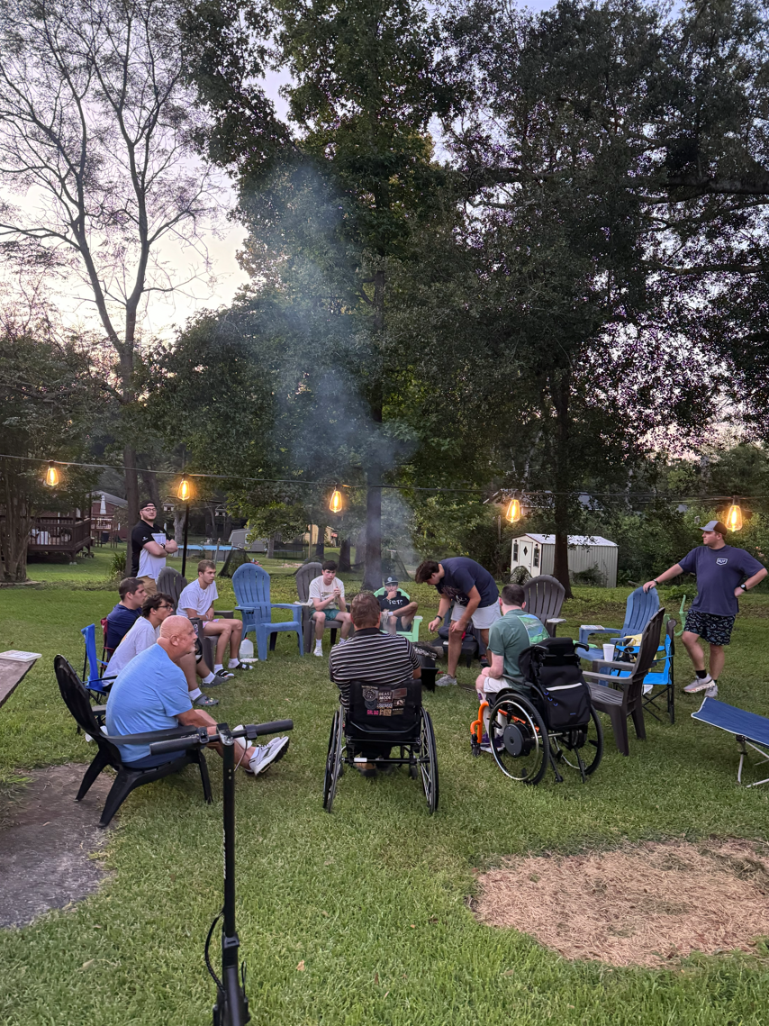 People gathered around a campfire on a lawn, some sitting in chairs, others standing or in wheelchairs, during dusk outdoors.