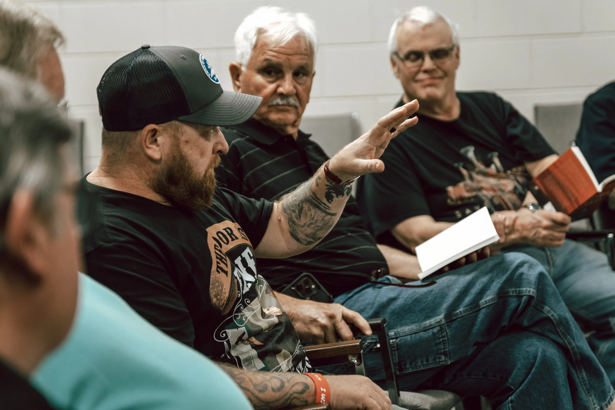 Group of men seated in a circle at a meeting or discussion, one man raising his hand while others hold notebooks.