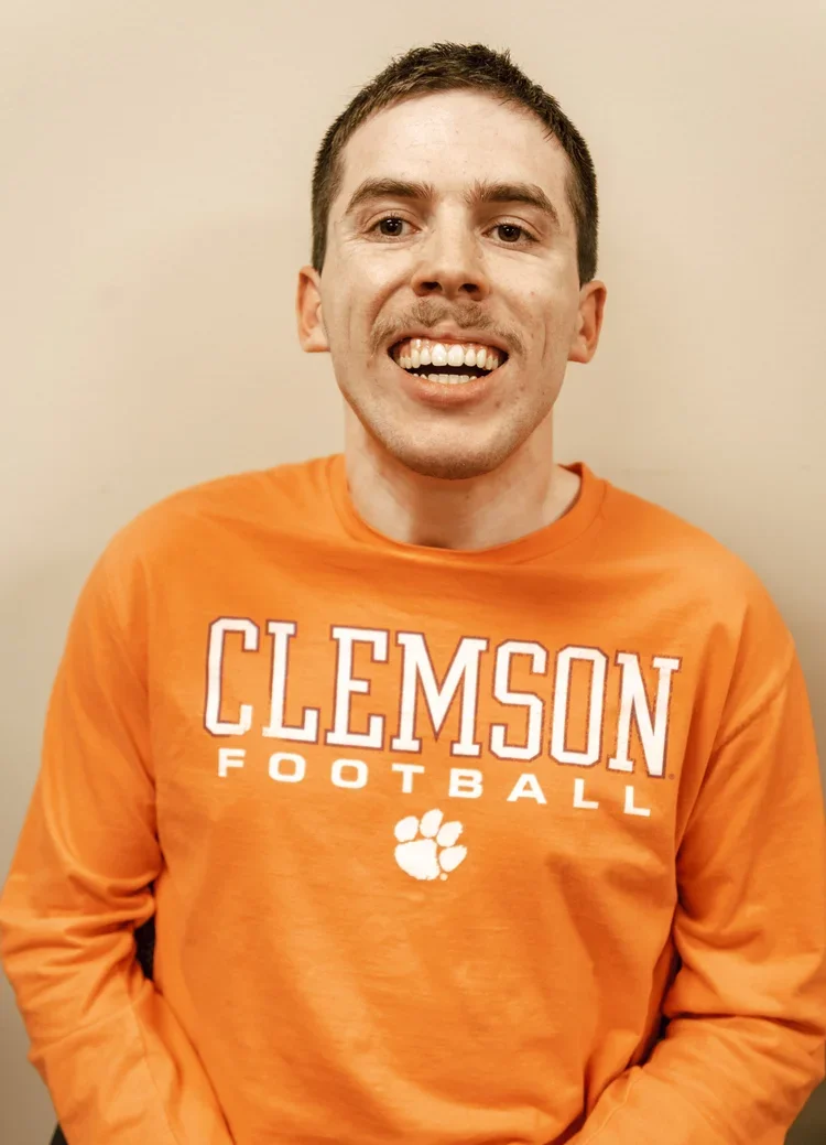 A young man smiling, wearing an orange Clemson Football sweatshirt with a paw print logo.