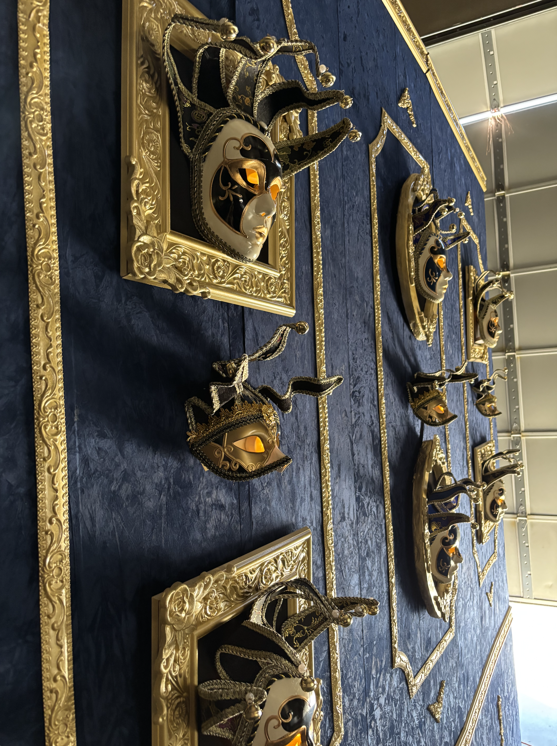 Multiple ornate Venetian masks with black, gold, and silver details, displayed on blue fabric with gold decorative borders. Baroque style theatre wall.
