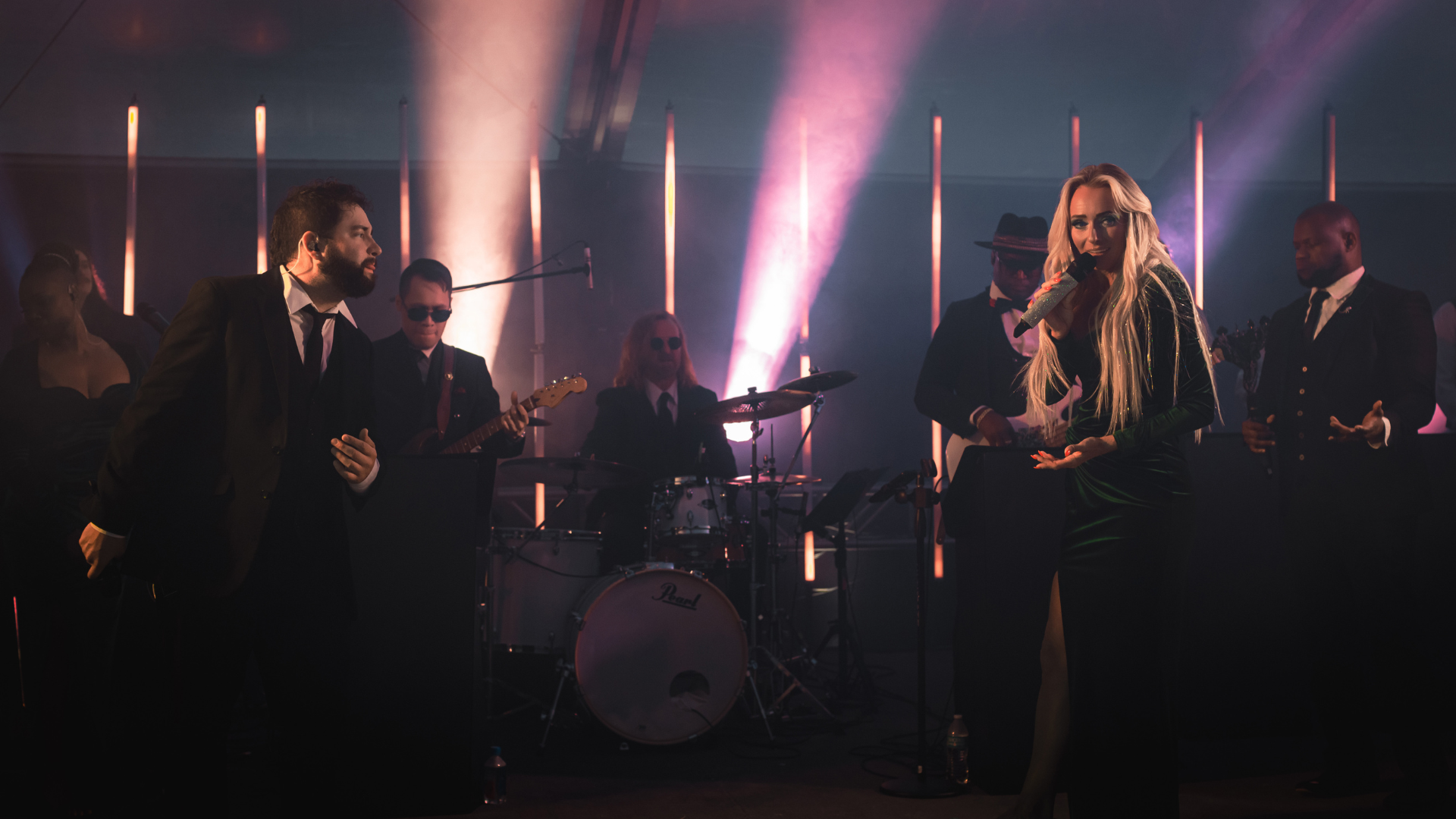 A live musical performance featuring a female singer with long blond hair, wearing a black dress, singing into a microphone with a glittering handle. She is surrounded by male and female band members dressed in formal black suits, playing instruments, on a stage with dramatic lighting including pink and purple spotlights.