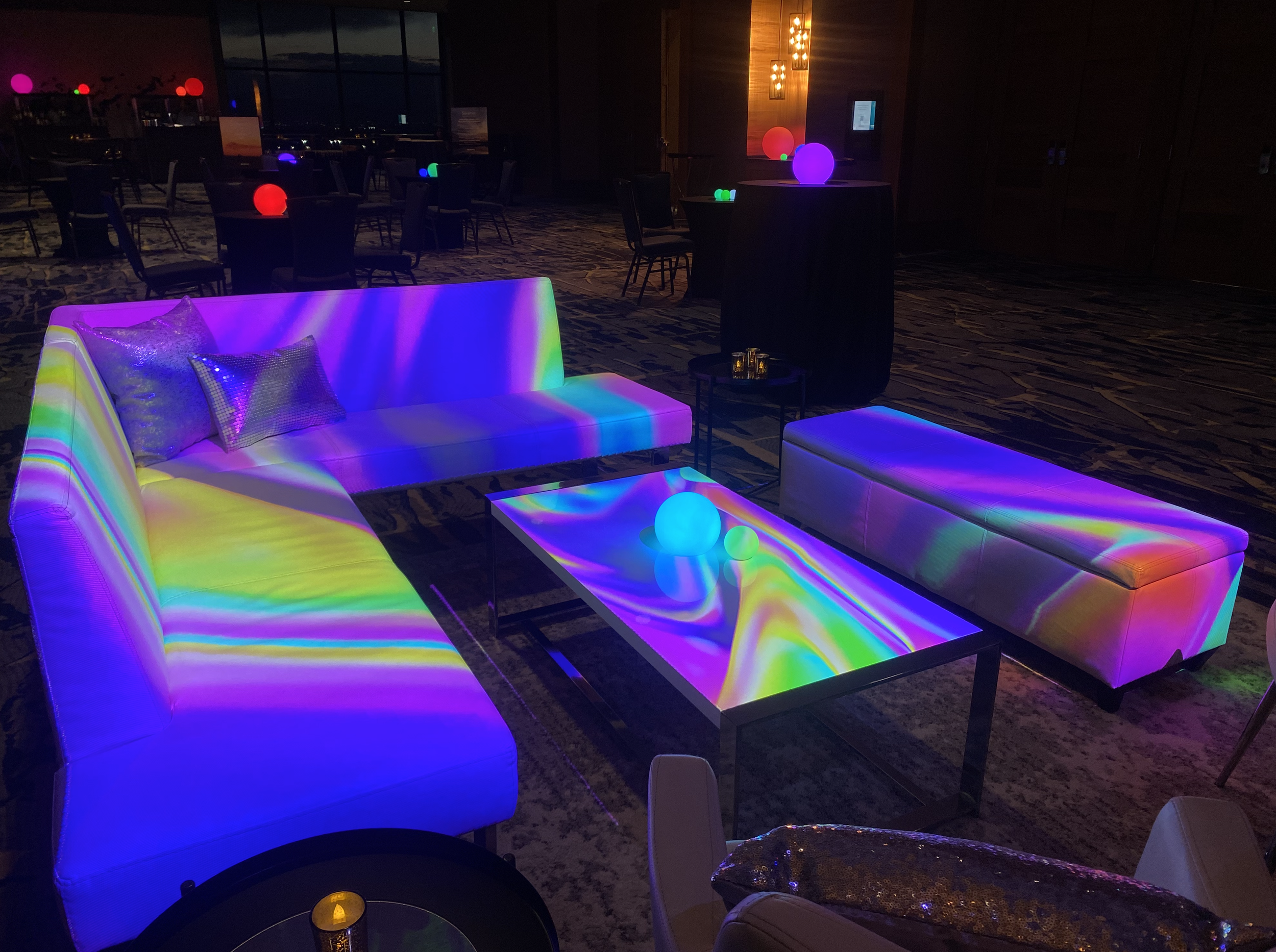 A modern lounge area illuminated with colorful, glowing lights that reflect on the furniture, including sofas and a table with spherical decorative lights, inside a dimly lit room with tables and chairs in the background.