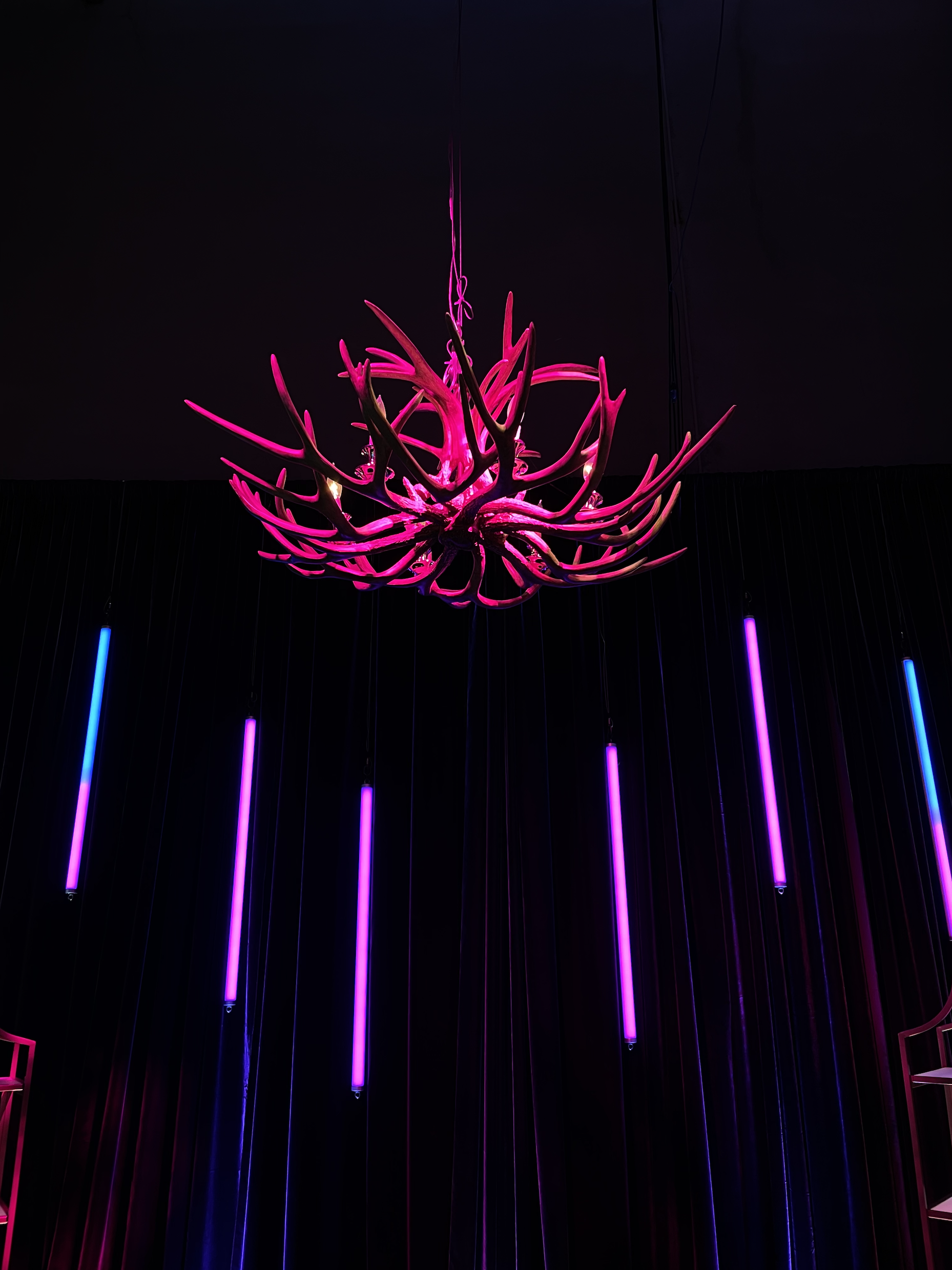 A pink chandelier with a modern, organic design hanging from a dark ceiling, illuminated by purple and blue neon lights in the background.