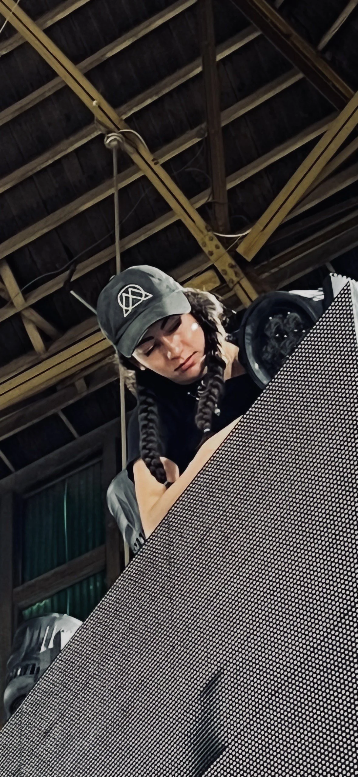 A woman with long braided hair, wearing a cap and a black shirt, looking at a container or object on a table in a rustic barn or warehouse with wooden beams overhead.