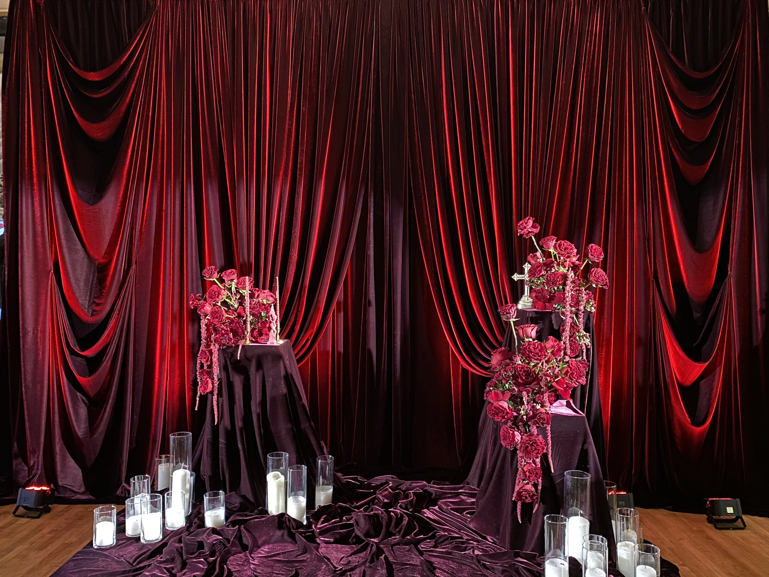Red velvet curtain backdrop with two tables decorated with pink and red flowers, with candles in glass holders on the floor.