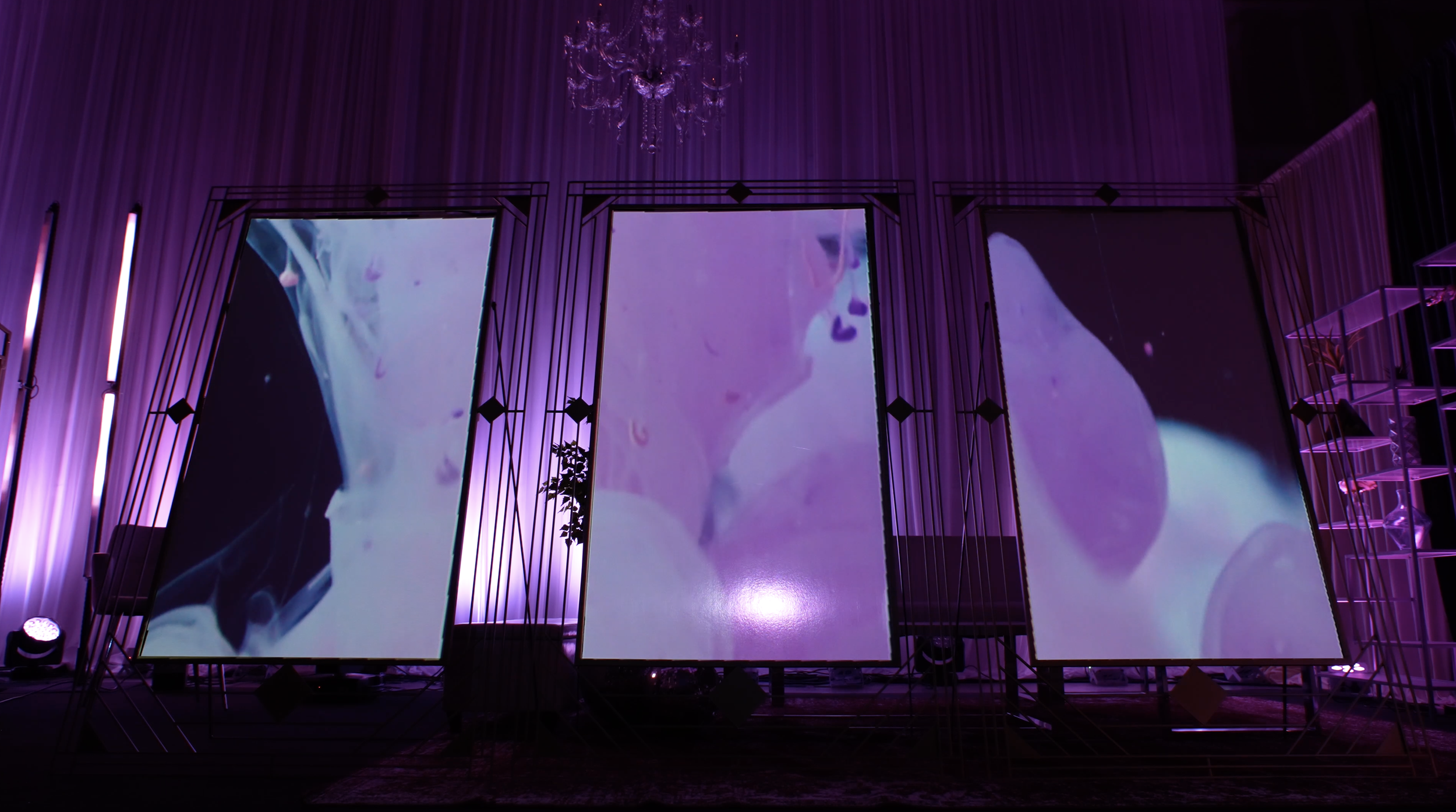 Three large vertical screens displaying abstract purple and pink imagery, surrounded by purple curtains and decorative lighting in a dimly lit room.