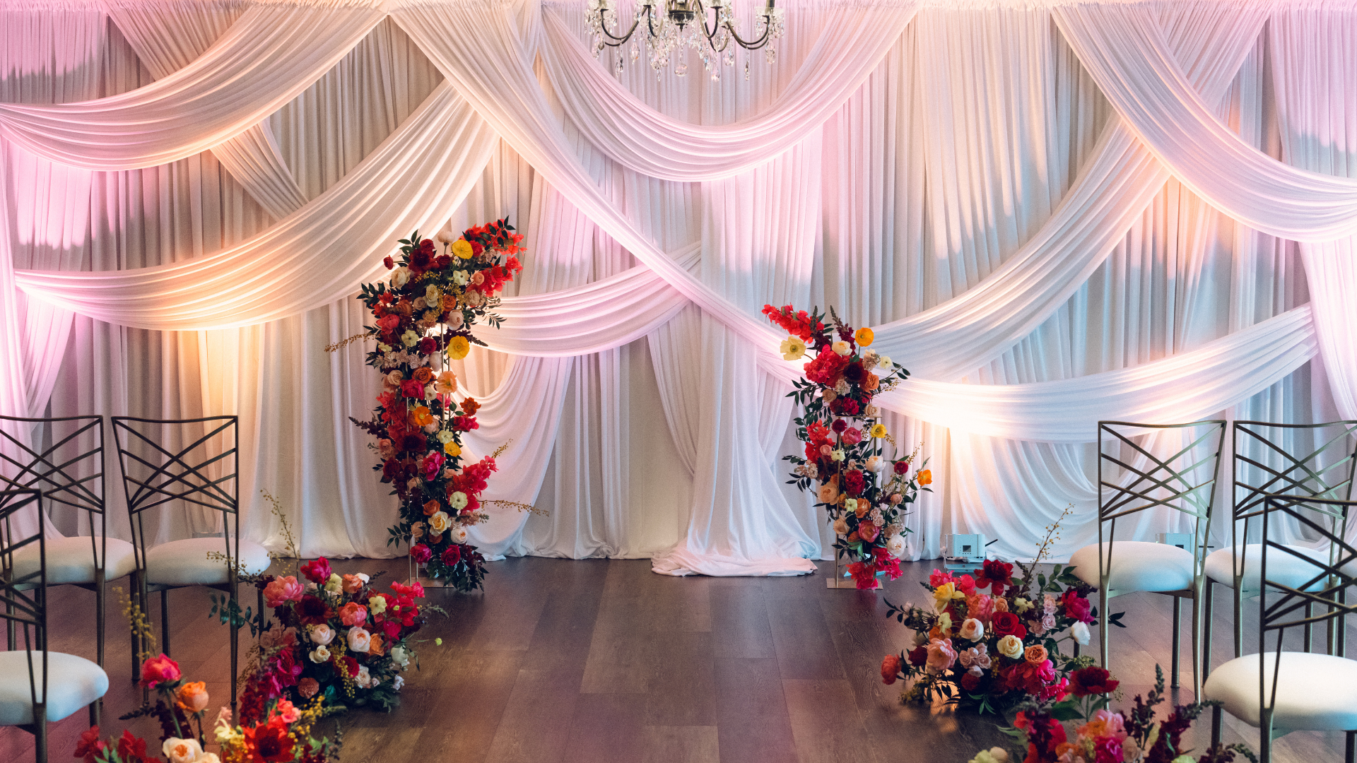 Wedding altar decorated with flowing white and pink drapes, and with red, pink, orange, and yellow floral arrangements on either side.