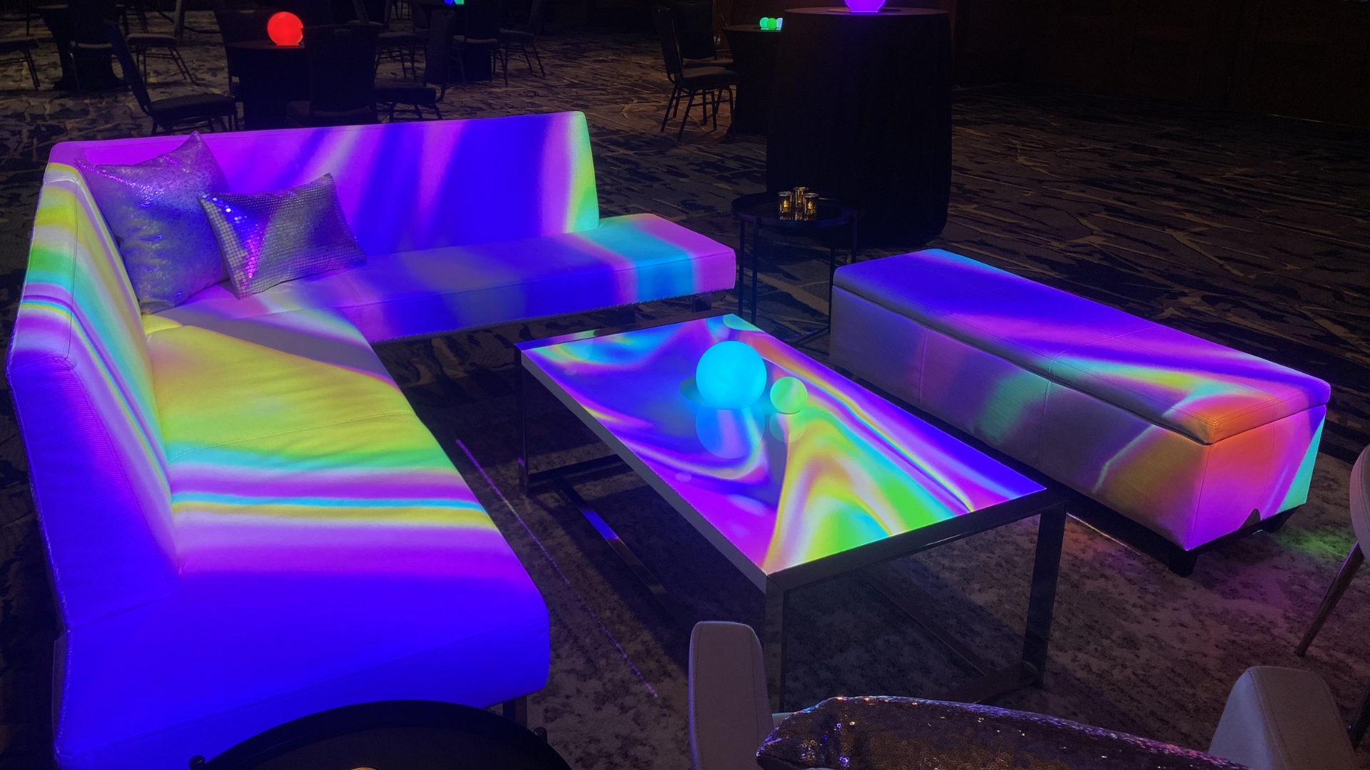 Colorful lounge area with holographic sofas, a coffee table with glowing orbs, and minimal lighting in a dim setting.