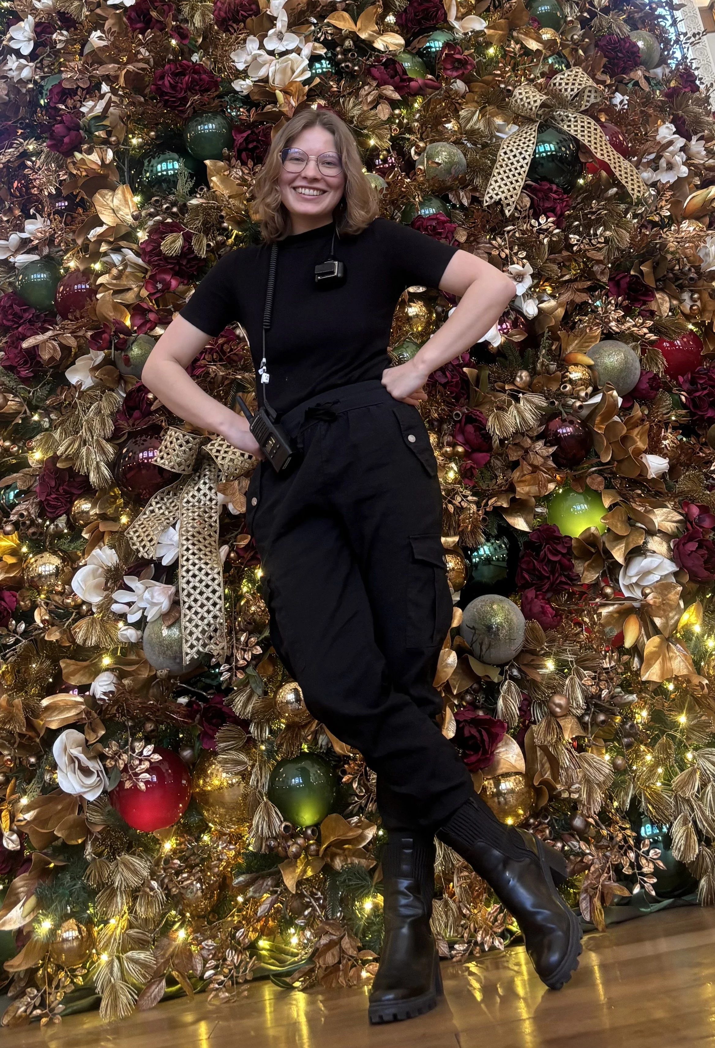 A woman with glasses and curly hair dressed in black, smiling and posing with hands on hips in front of a decorated Christmas tree with gold, red, green, and white ornaments and lights.
