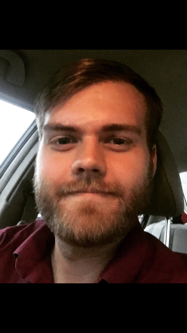 Close-up of a man with light brown hair and a beard, sitting in a car. He is wearing a dark red shirt and looking at the camera with a neutral expression.