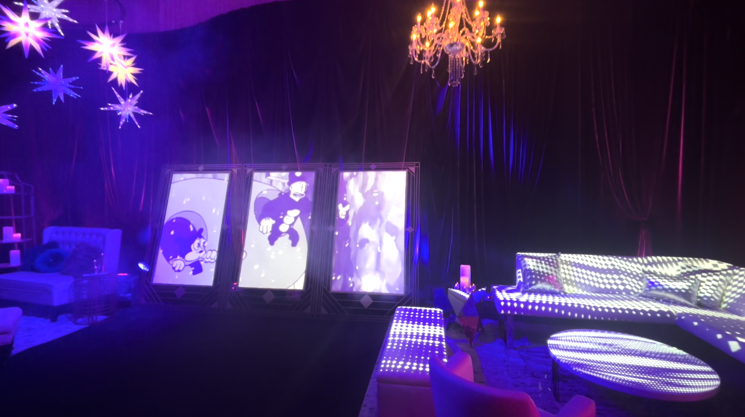 A room decorated with old cartoons on large screens, illuminated with purple and blue lights, featuring a chandelier, star-shaped ceiling lights, a white sofa with lights projected on it, and purple accents for a themed party or event.