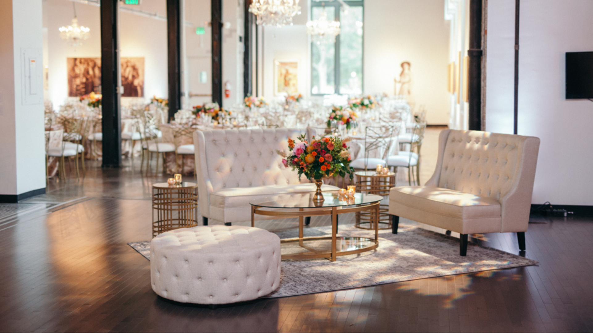 Elegant event space with white tufted sofas and a central glass table with a floral arrangement. The background shows round tables with floral centerpieces and gold chairs, chandeliers, and artwork on the walls, all illuminated by natural light.