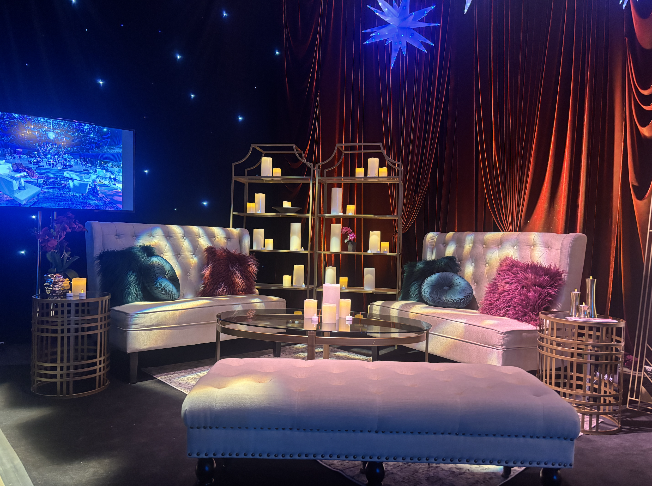 A cozy living room with two tufted sofas, colorful throw pillows, candles, a large TV, and a decorative shelving unit, illuminated by soft lighting, with a star-shaped ornament hanging from the ceiling.