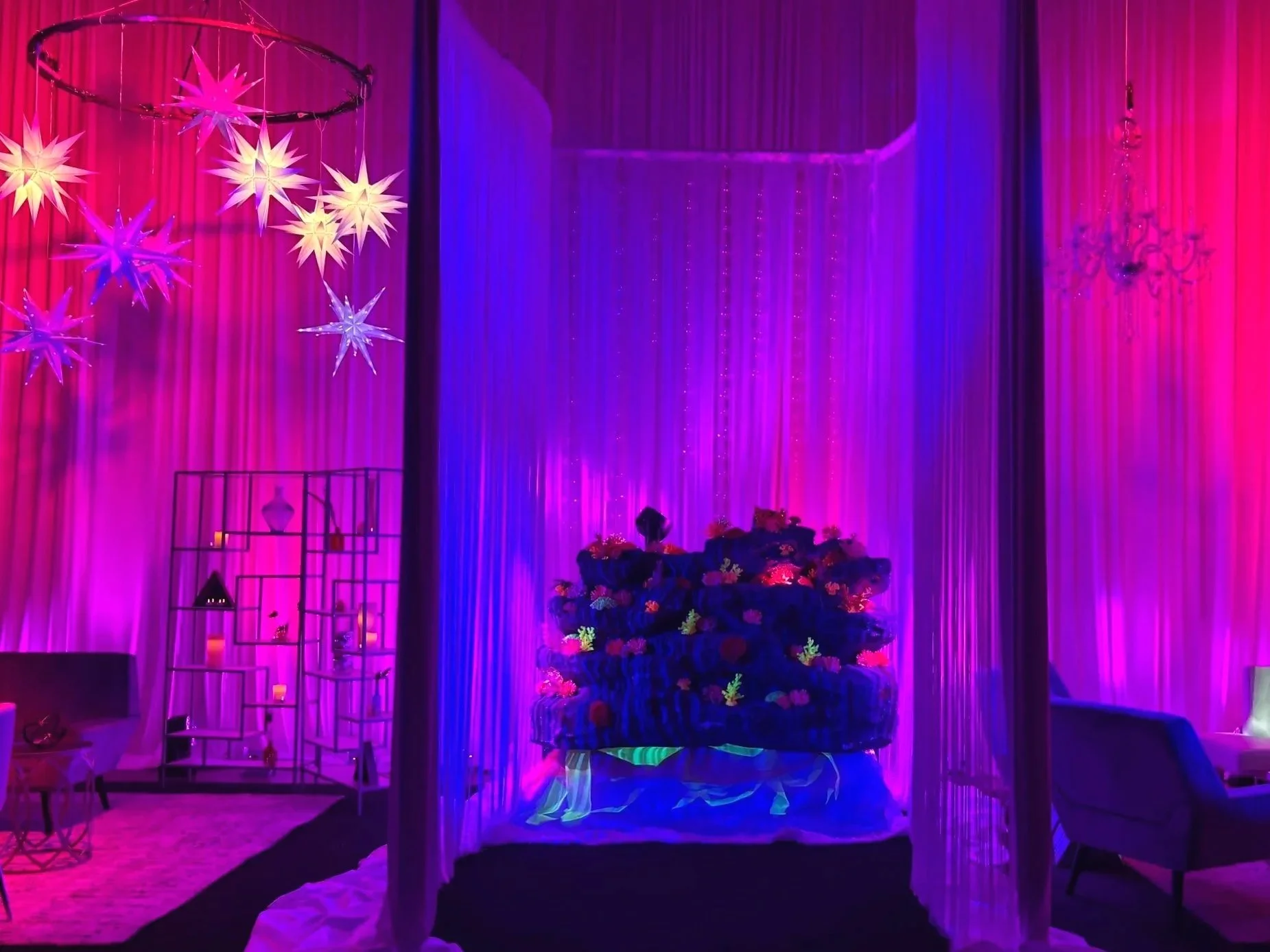 Colorful decorated film studio with hanging star-shaped lights, purple curtains, a floral arrangement on a bed, and various furniture pieces illuminated with vibrant purple, pink, and blue lighting.