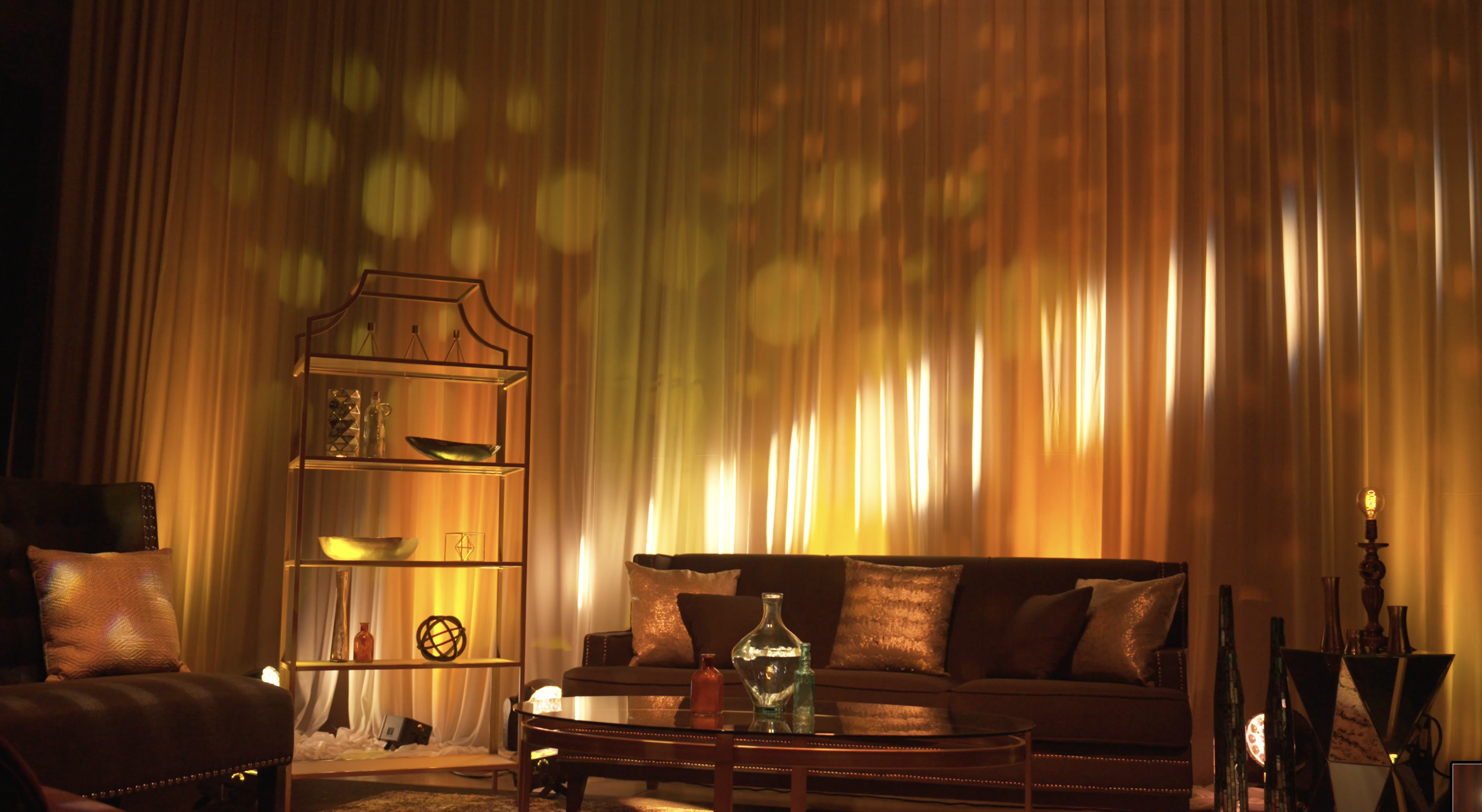 A cozy living room with a dark brown leather sofa and a black and white armchair, decorated with shiny pillows. A round glass coffee table with decorative bottles and a vase sits in front of the sofa. Behind, a textured curtain in warm yellow and orange tones creates a glowing background, with ambient lighting casting a soft, warm glow across the room.