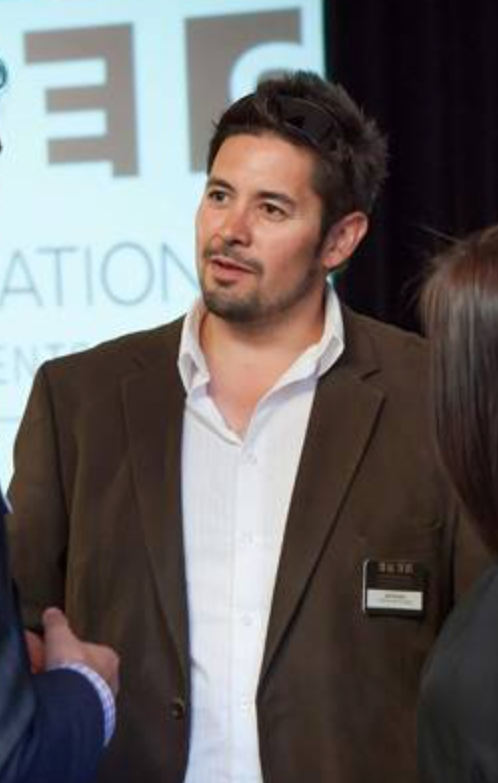 Man in brown suit jacket and white shirt speaking with two other people at a professional event.