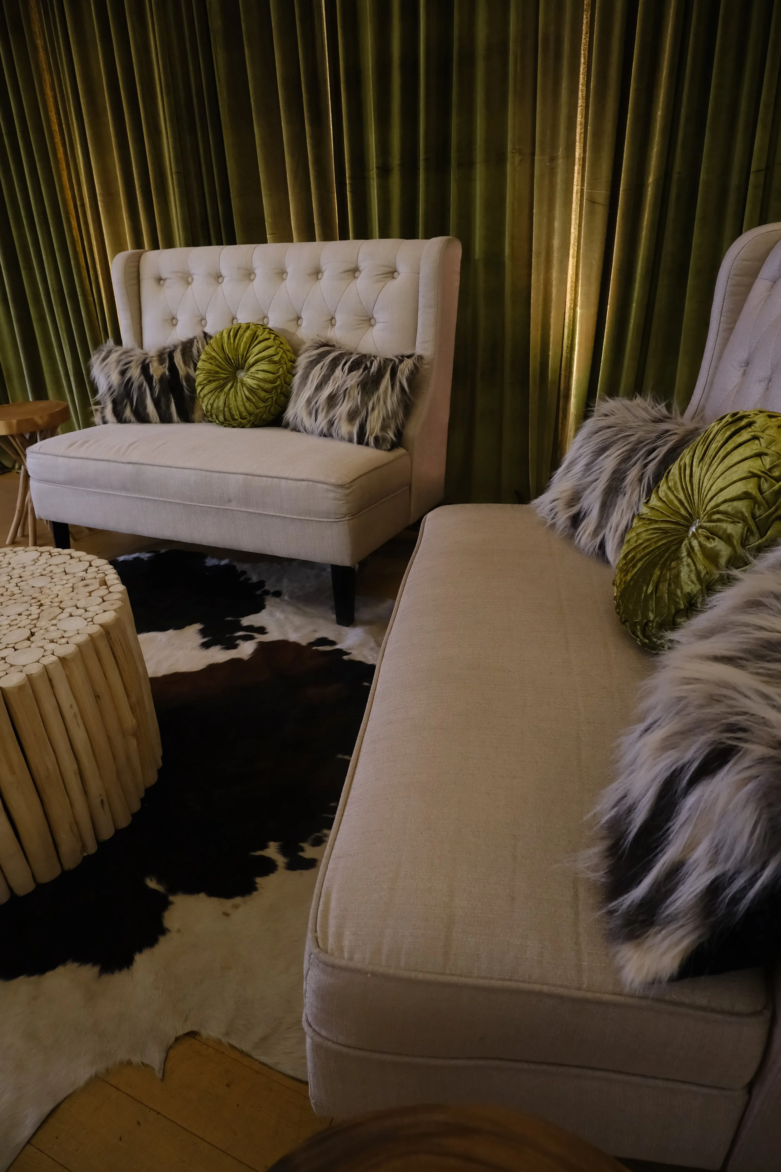 Living room with beige upholstered armchair and a matching sofa, decorated with green round pillows and faux fur pillows, green curtains, wooden side table, and cowhide rug.
