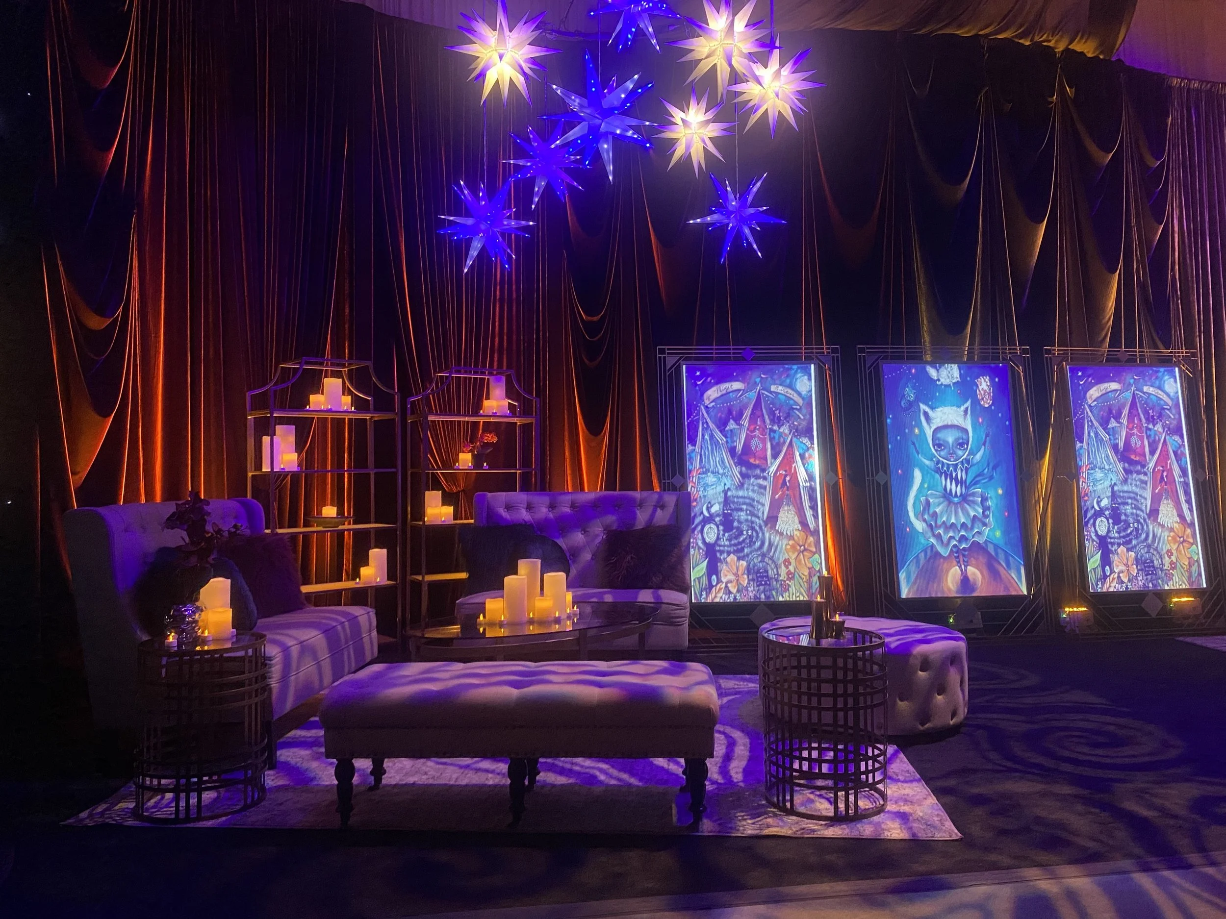 A cozy lounge area decorated with purple sofas, ottomans, and candlelit tables. Behind, there are tall shelves with candles, dark curtains with orange accents, and vibrant, colorful artworks featuring whimsical, fantasy-themed designs. Hanging star-shaped lights cast a warm glow over the scene.