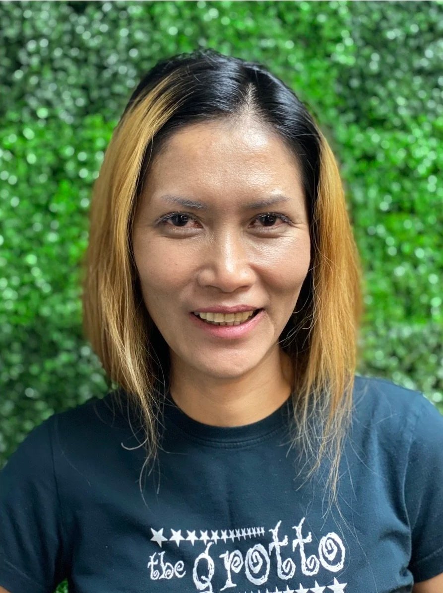 A woman with shoulder-length hair, balayage highlights, and a dark shirt smiling in front of a green leafy background.