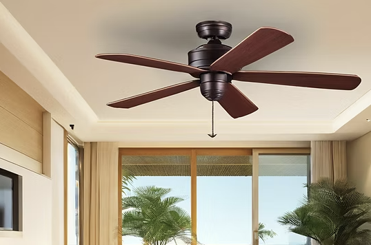 Ceiling fan with wooden blades in a room with large windows and tropical plants outside.
